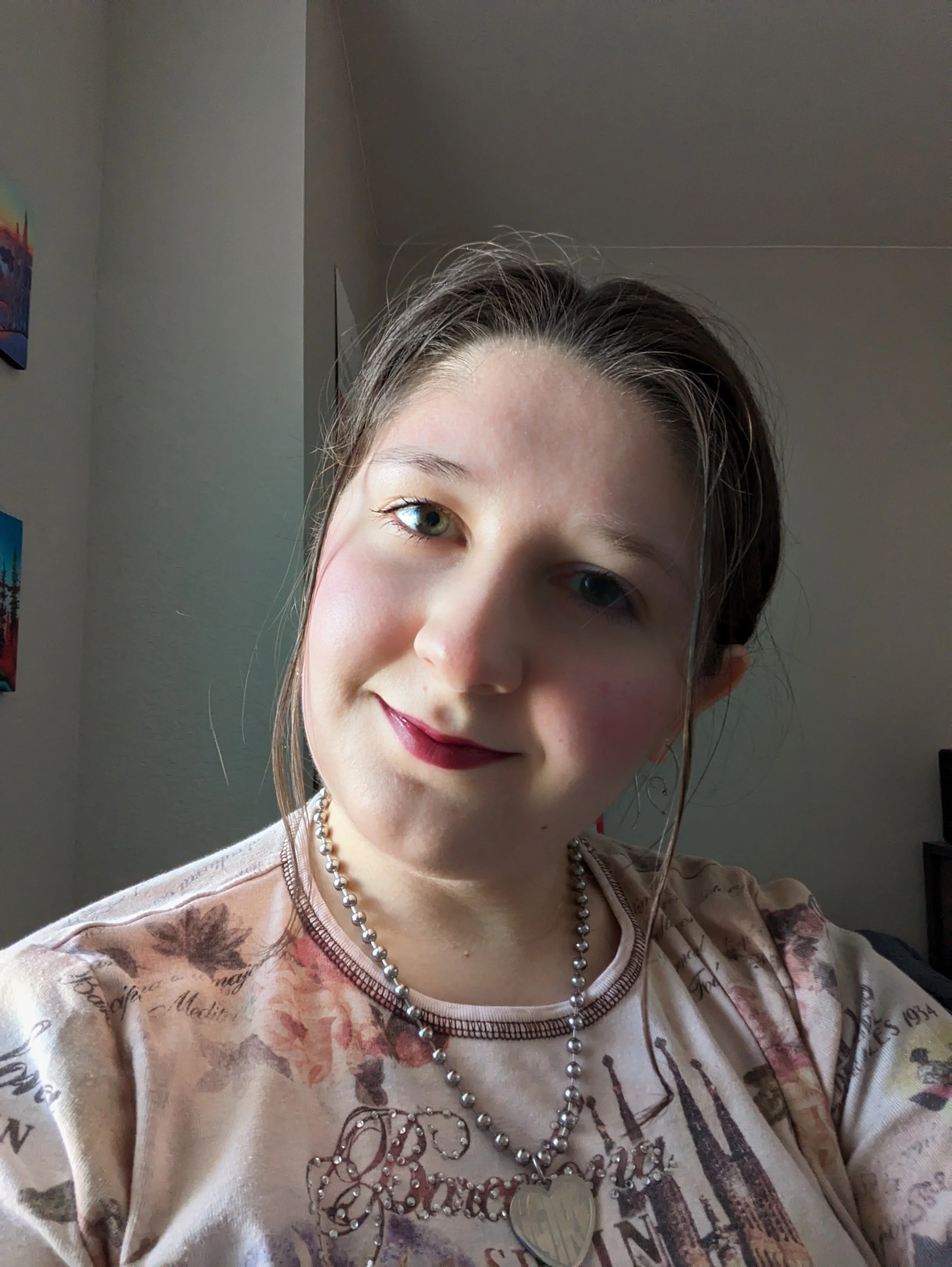 A young woman with brown hair, wearing a pearl necklace with a heart-shaped pendant, smiling at the camera in a room with light-colored walls and artwork.