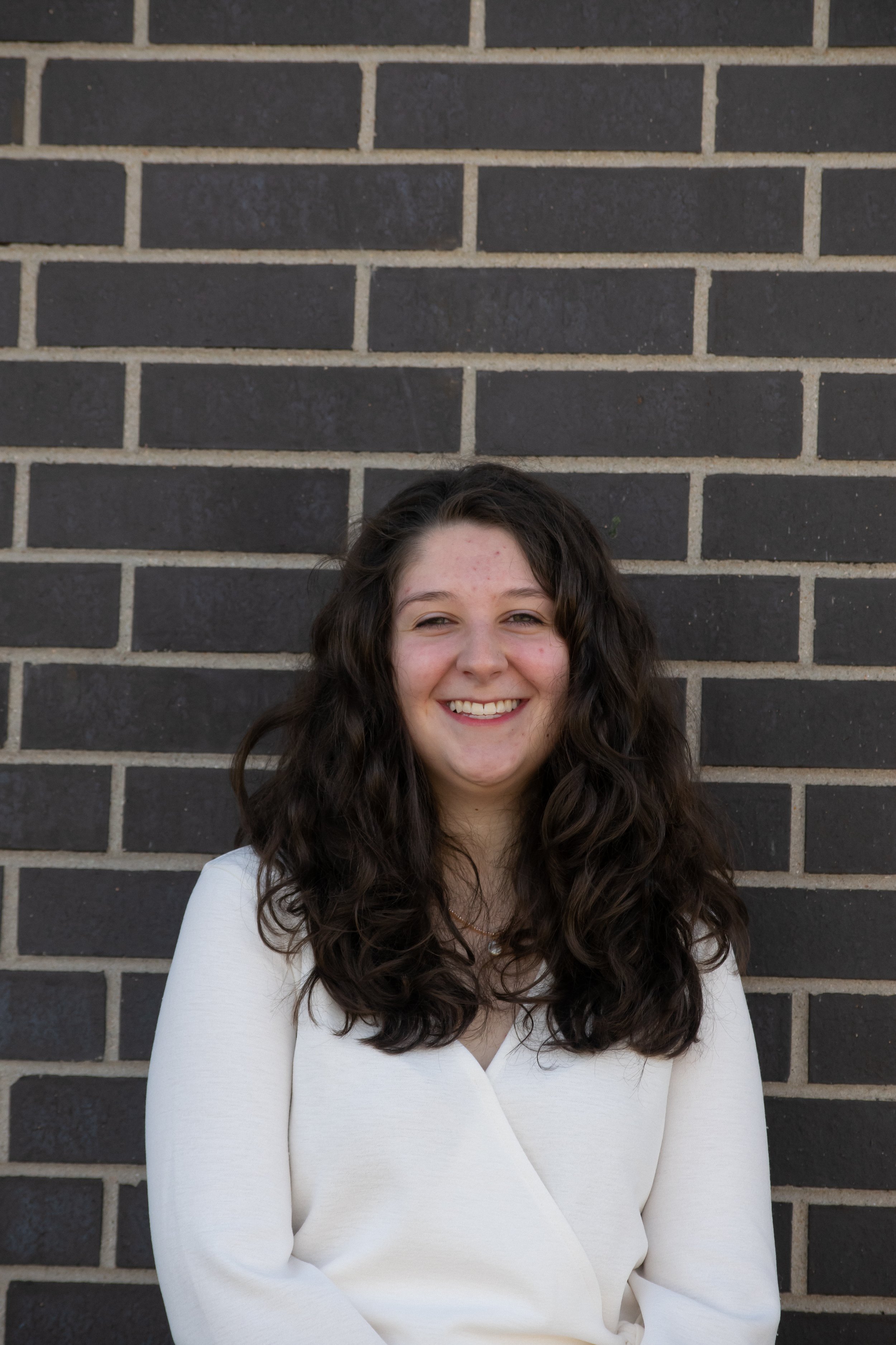 A young woman with long curly brown hair, wearing a white top, smiling and standing against a dark brick wall.