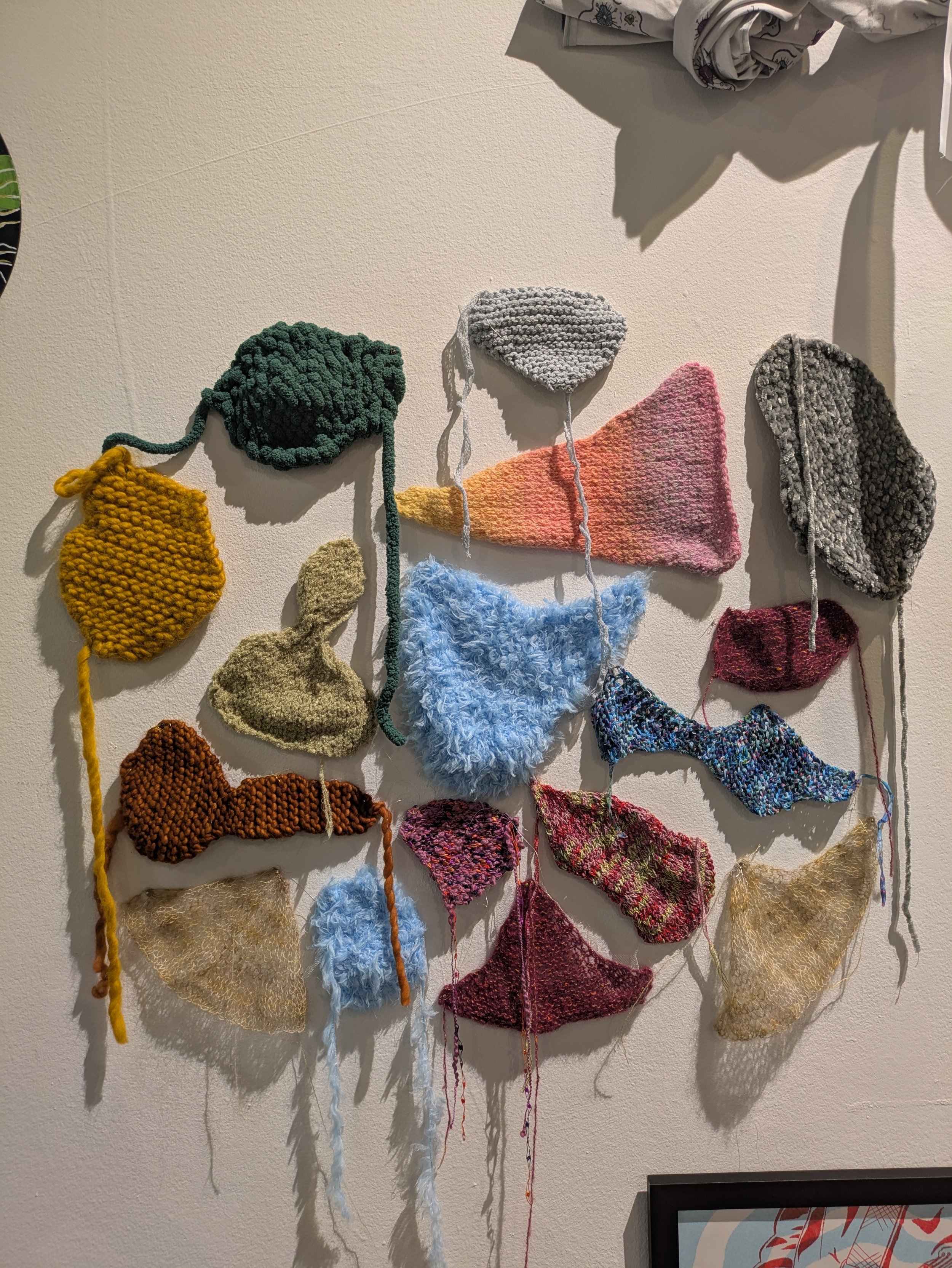 A wall display of various colorful knitted shapes of different textures and sizes, resembling abstract objects or clouds.
