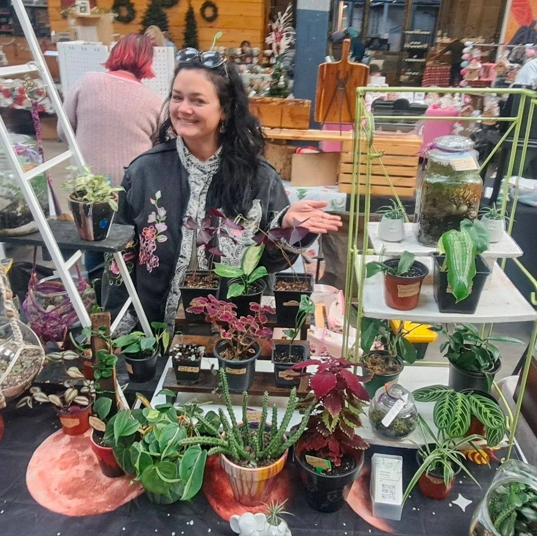 Smiling woman at a plant and gardening display with various potted plants, succulents, and decorative terrariums on tables