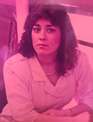 Woman with curly dark hair wearing a light-colored blouse and pearl necklace, sitting with arms crossed, with a pink hue filter.