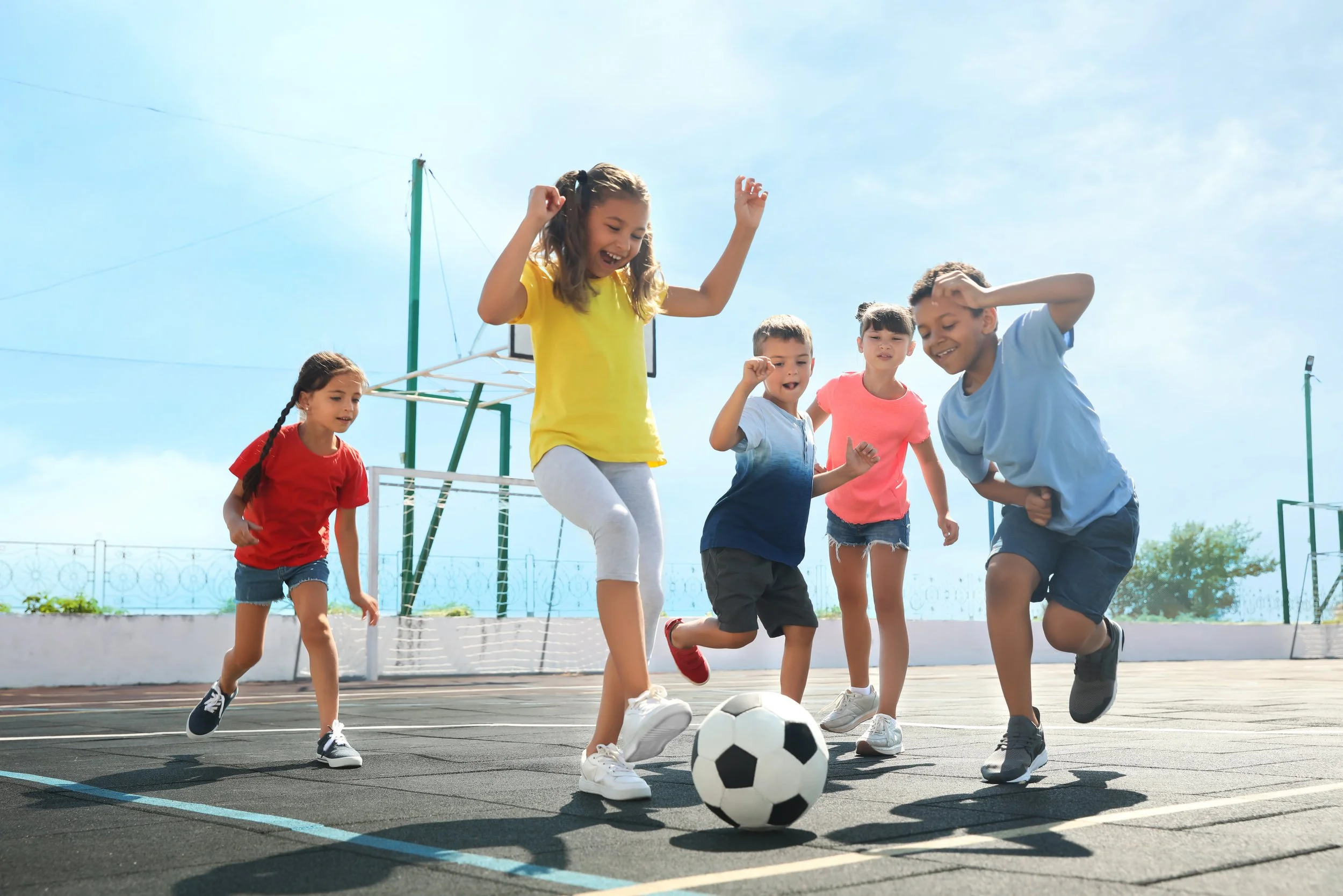 Children playing soccer on an outdoor court, joyful and energetic, under a clear blue sky.