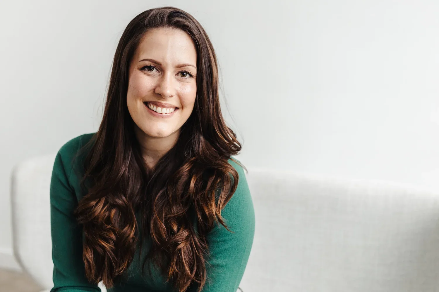A woman with long, wavy brown hair, wearing a green shirt, smiling while sitting indoors against a light-colored wall.