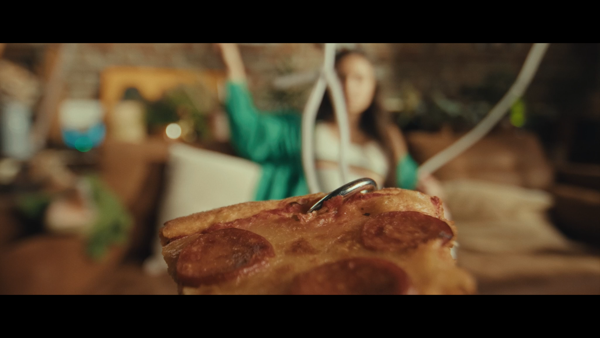 Une part de pizza au pepperoni en premier plan, floue d'arrière-plan, où une femme est assise dans un environnement intérieur avec des décorations en bois et des plantes.