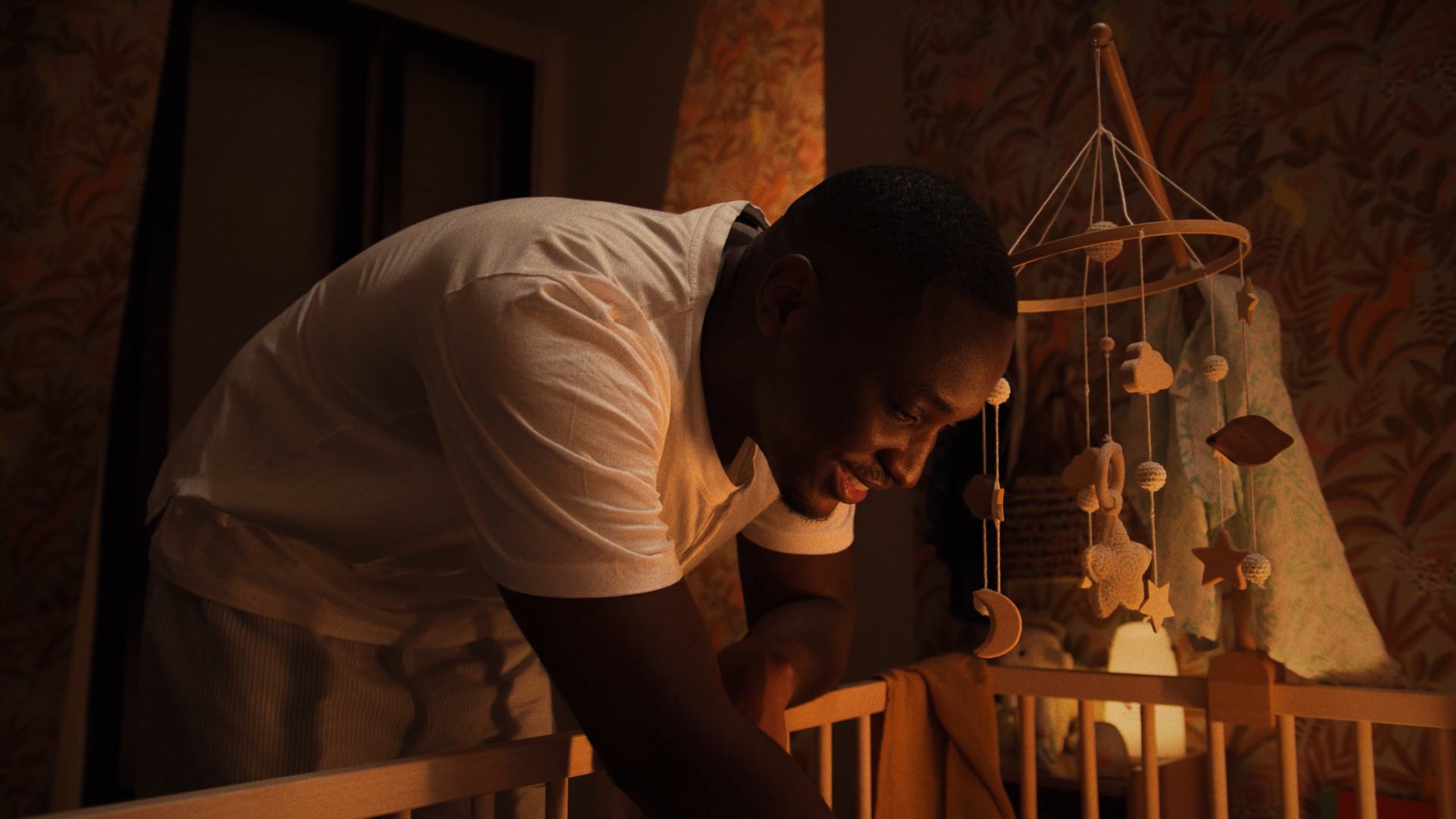 Un homme souriant dans une chambre à la lumière tamisée, près d'un lit, avec une décoration de mobile suspendu avec des étoiles, des lunes et des formes de nuages.