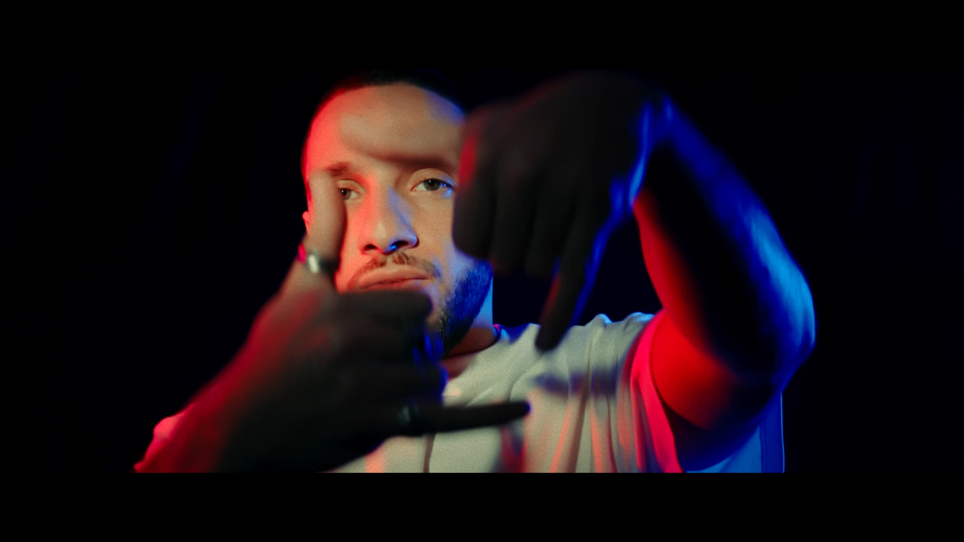 Un homme avec une barbe regarde l'objectif, il porte un t-shirt blanc et des gants noirs. Il forme un cadre avec ses mains, éclairage en rouge et bleu sur un fond noir.