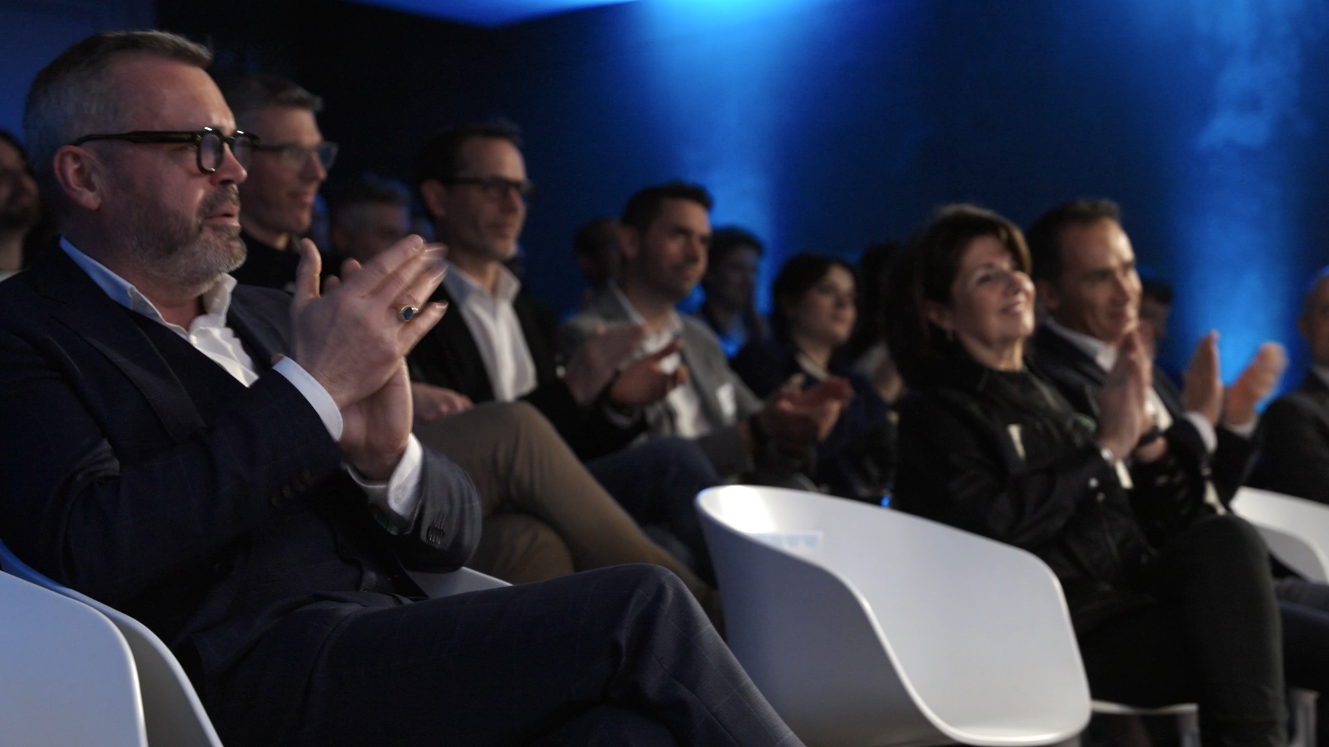 Groupe de personnes assises et applaudissant lors d'une conférence ou d'un événement, dans une salle sombre avec un éclairage bleu.