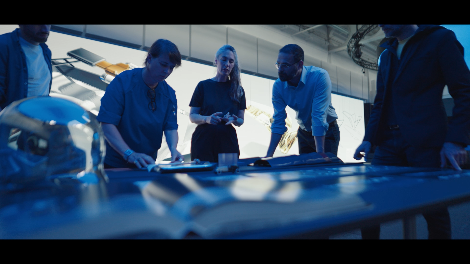 Groupe de cinq personnes discutant autour d'une table avec des plans et une maquette d'avion dans un environnement moderne et technologique.
