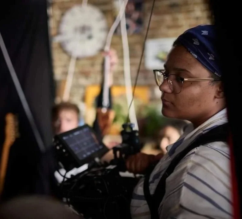 Une personne portant des lunettes, un bandana bleu et un vêtement rayé travaille avec un équipement électronique dans un environnement intérieur avec un mur en briques.