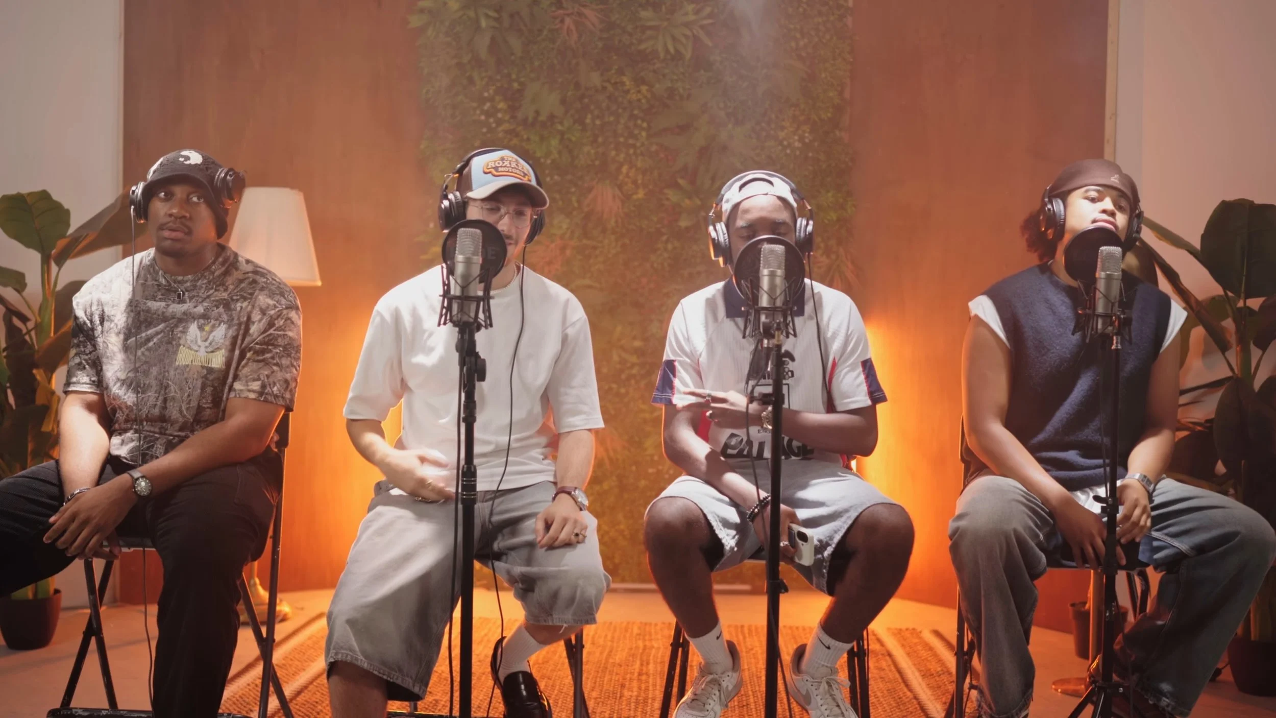 Quatre jeunes hommes assis dans un studio d'enregistrement, portant des écouteurs et parlant dans des micros, avec un décor chaleureux et végétal en fond.