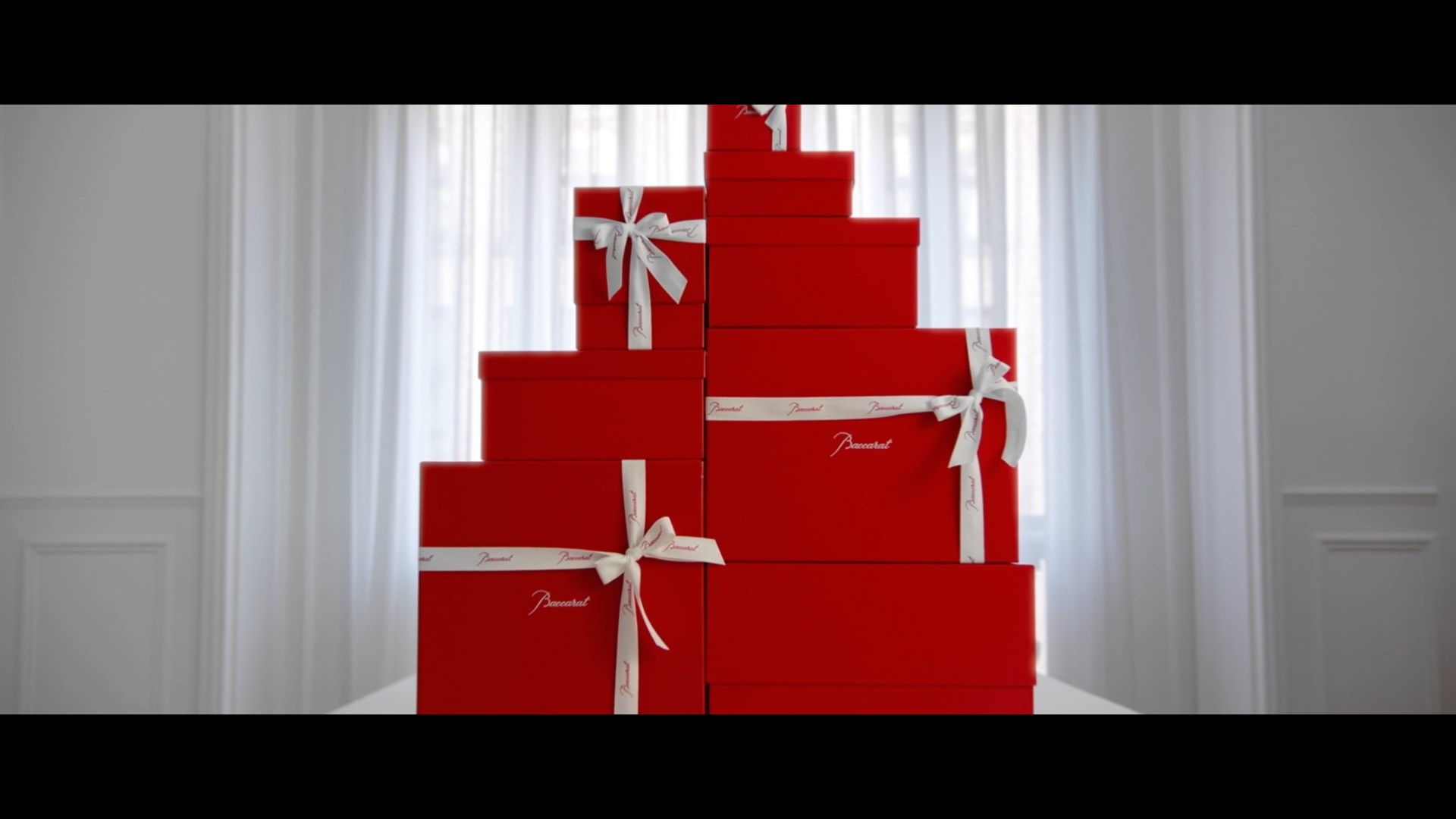 Piles de boîtes rouges de différentes tailles, décorées avec des rubans blancs avec le mot 'Baccarat', empilées en forme de sapin de Noël, dans une pièce lumineuse avec des rideaux blancs.