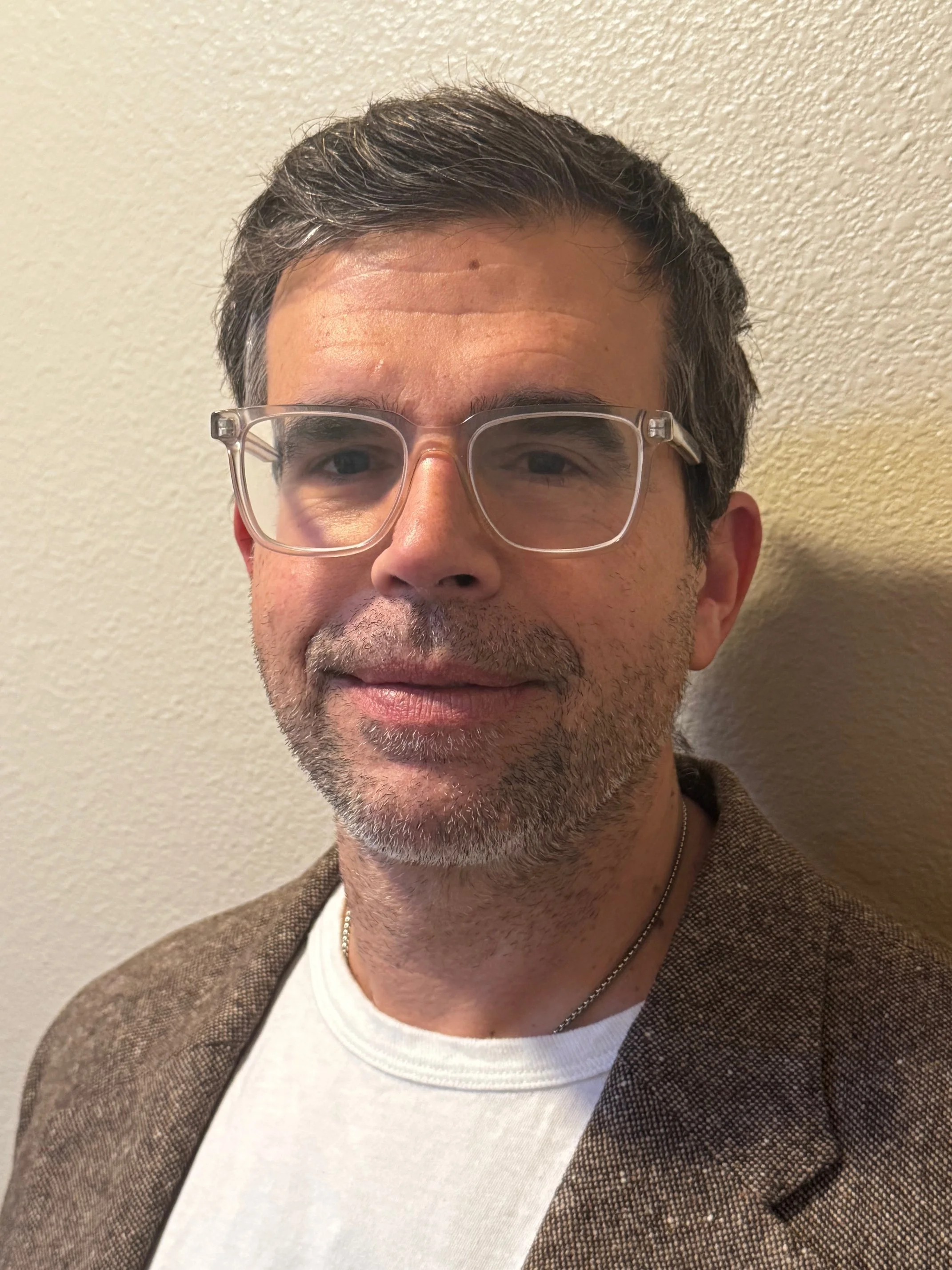A middle-aged man with dark hair and glasses smiling, wearing a white shirt and brown blazer, standing against a beige wall.