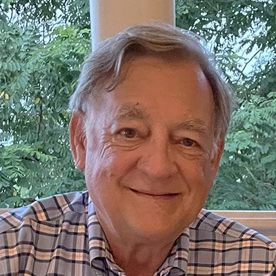 Portrait of a middle-aged man with gray hair smiling, wearing a plaid shirt, sitting indoors near a window with trees outside.