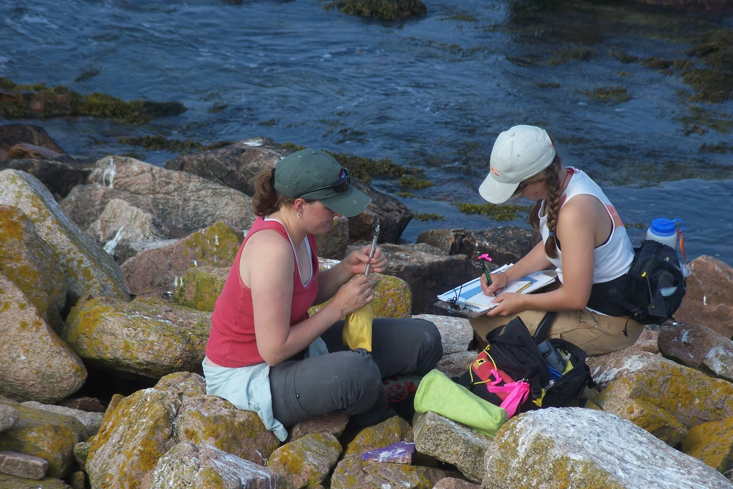 Two women sitting on rocks near a body of water, recording data or taking notes, engaged in outdoor research or survey activity.