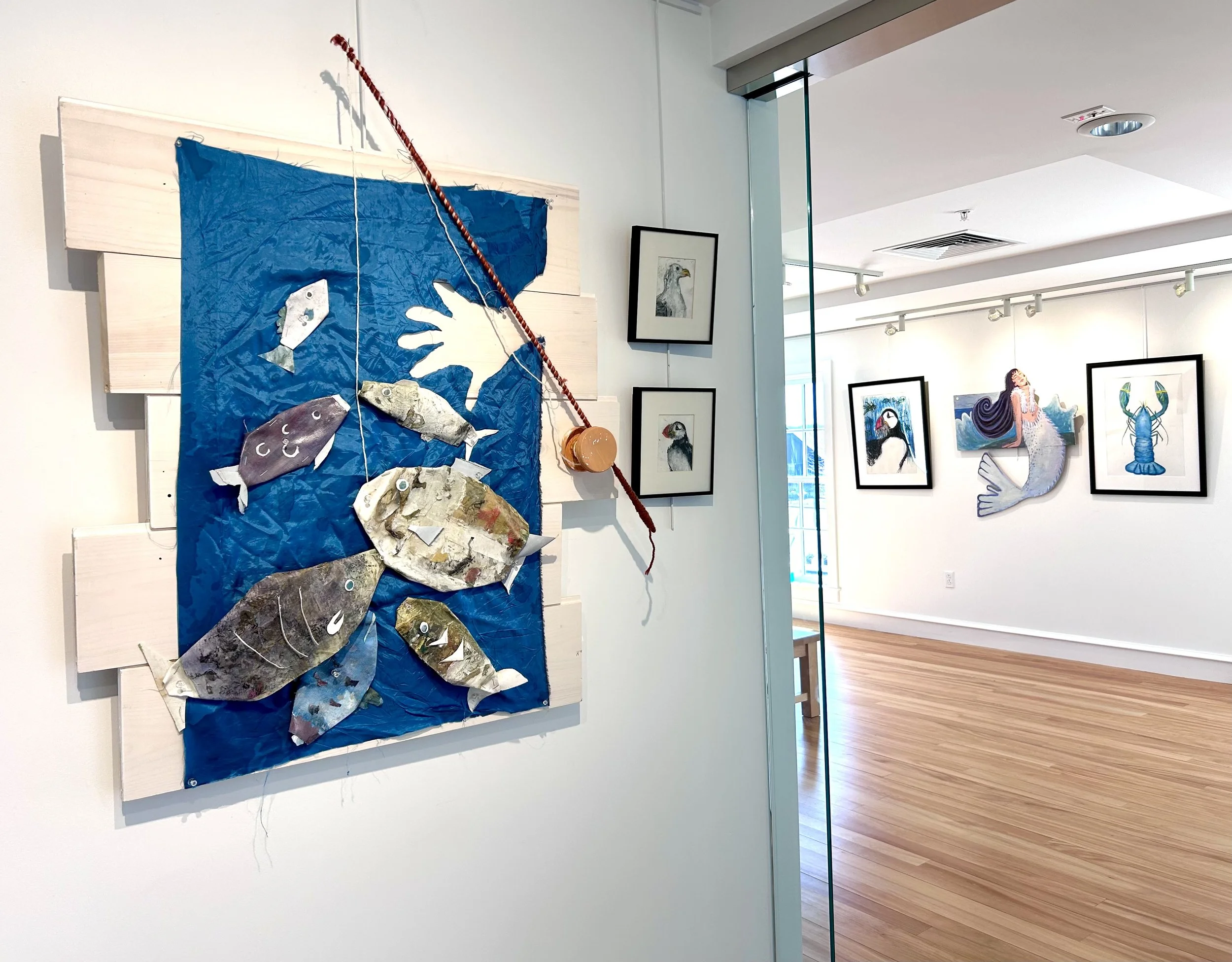Art gallery with whale and aquatic animal themed artwork on white walls, including framed drawings and a large mixed-media piece with fish and hands, and a wooden floor.
