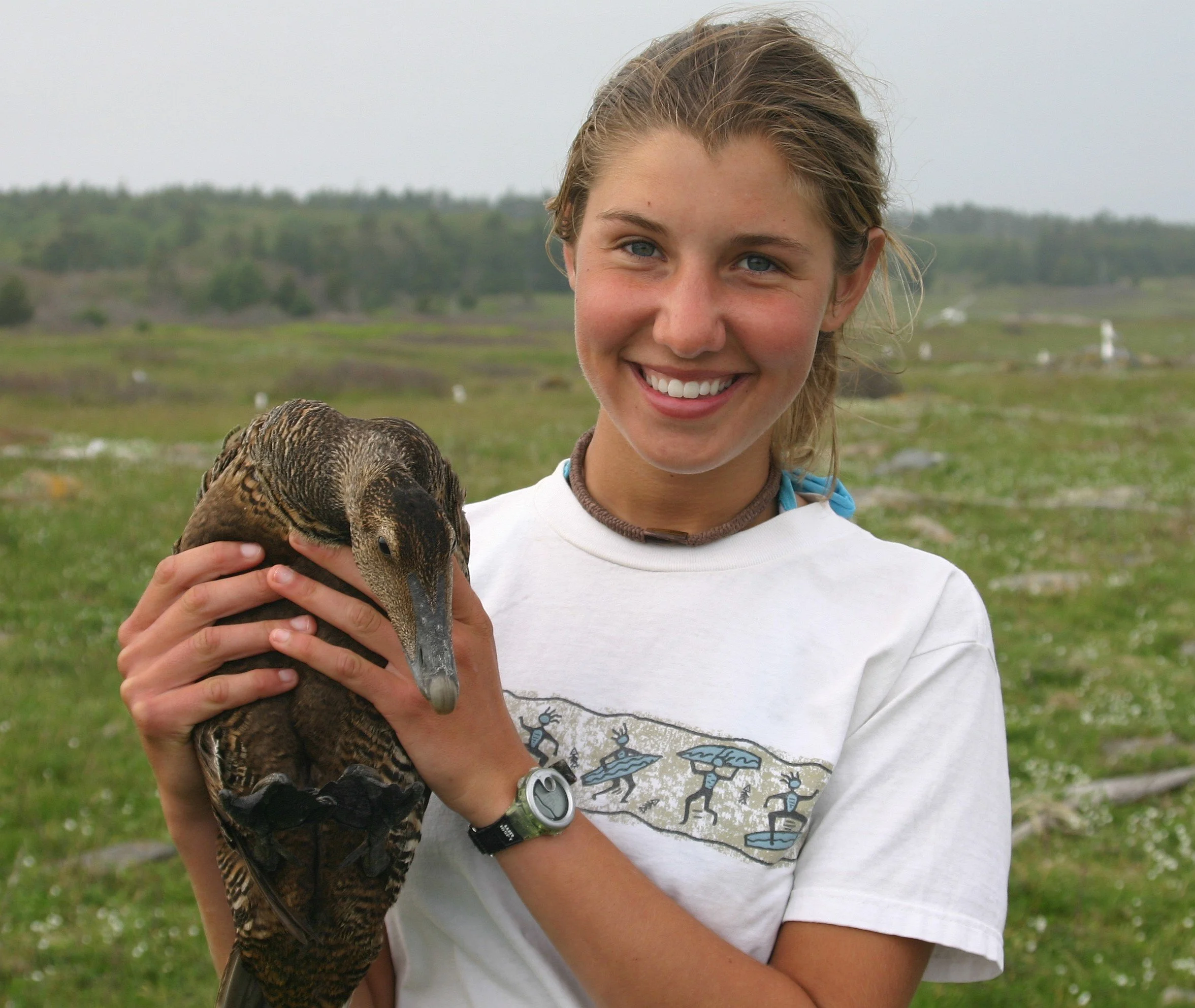 A smiling young woman holding a brown duck outdoors on a grassy field with a blurred natural background.