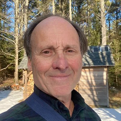 Man with short hair and a slight smile standing outdoors in front of trees and a wooden shed