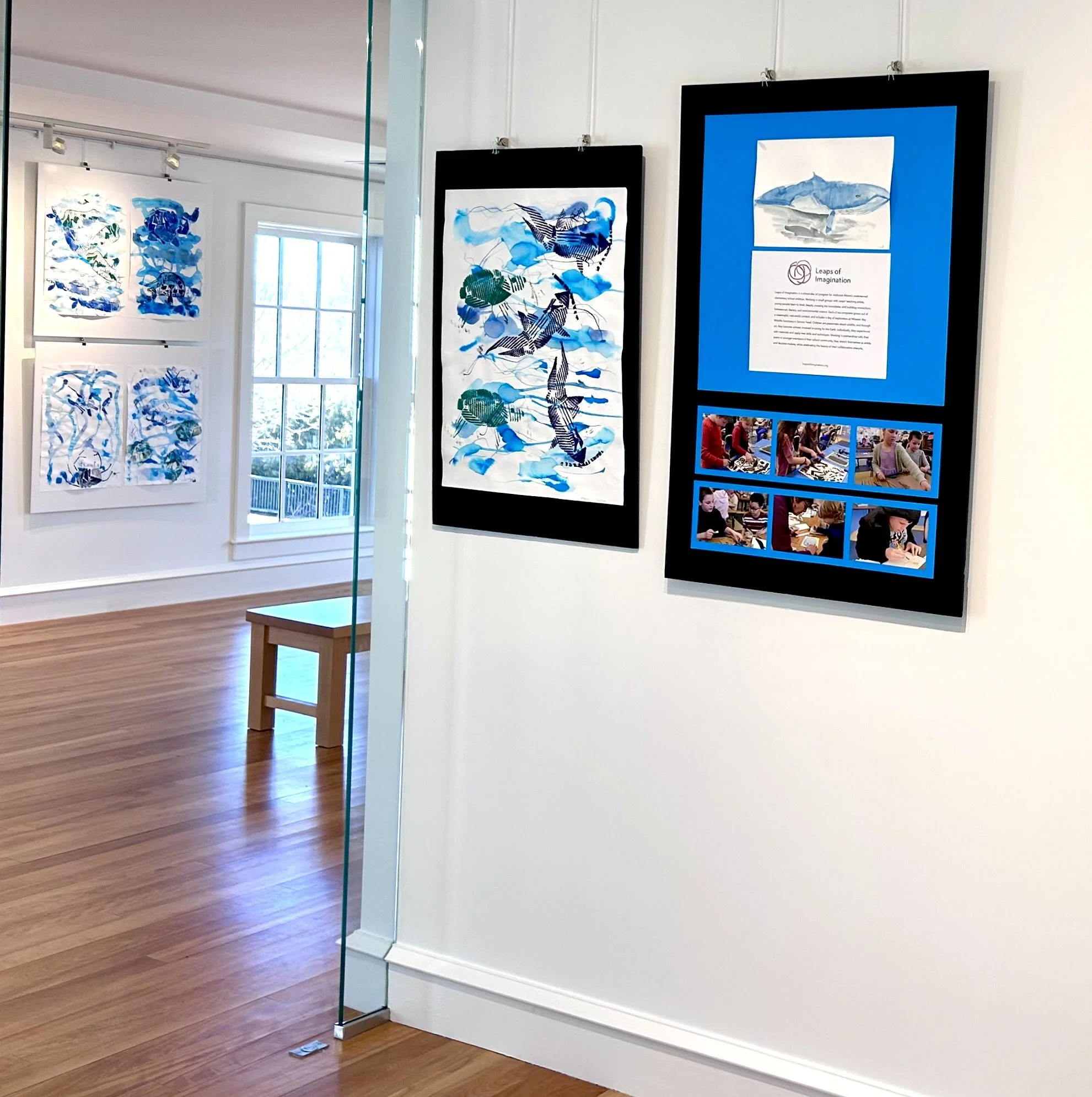 Art gallery displaying various framed abstract artworks with shades of blue and green, and some photographs of people working on art projects.