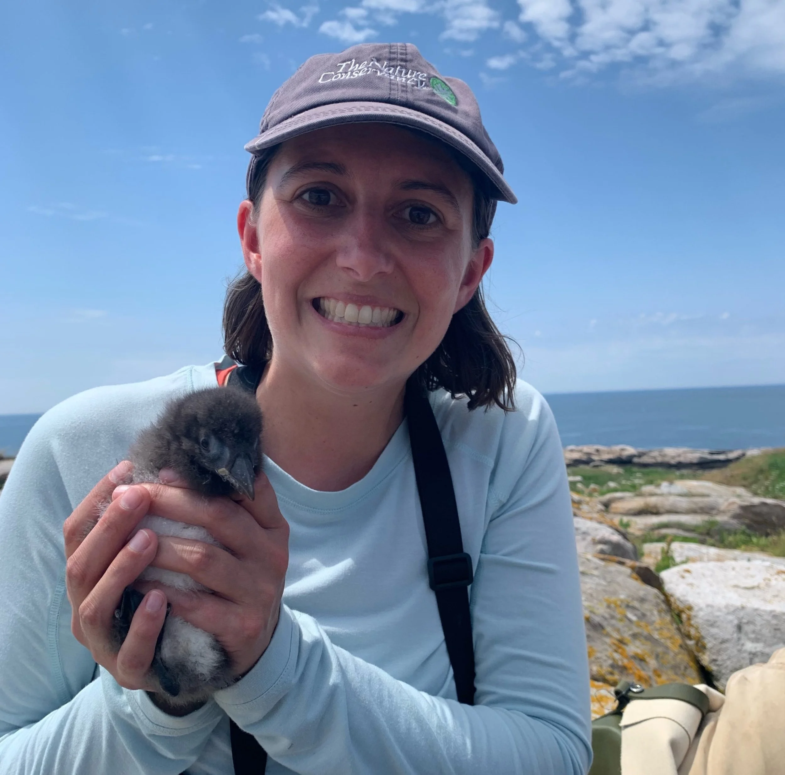 A woman smiling and holding a fluffy baby penguin near a rocky coastline with the ocean in the background.