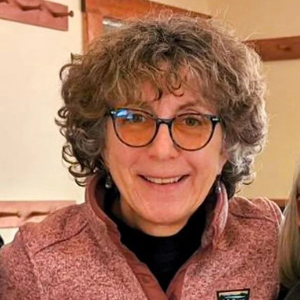 A woman with curly gray hair and glasses smiling for the camera indoors.