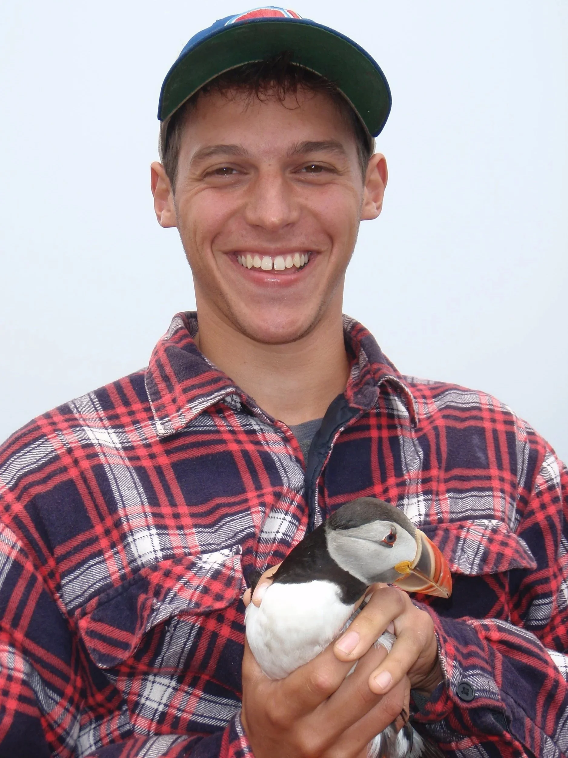 A young man smiling and holding a puffin bird in his hands, wearing a red and black plaid shirt and a baseball cap with a logo.
