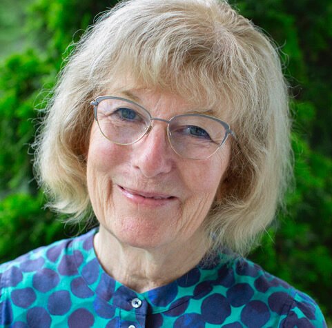 A woman with blonde hair and glasses smiling outdoors with a green leafy background.