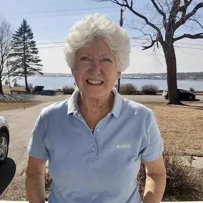 Older woman with white hair smiling outdoors near water, trees, and a park bench.