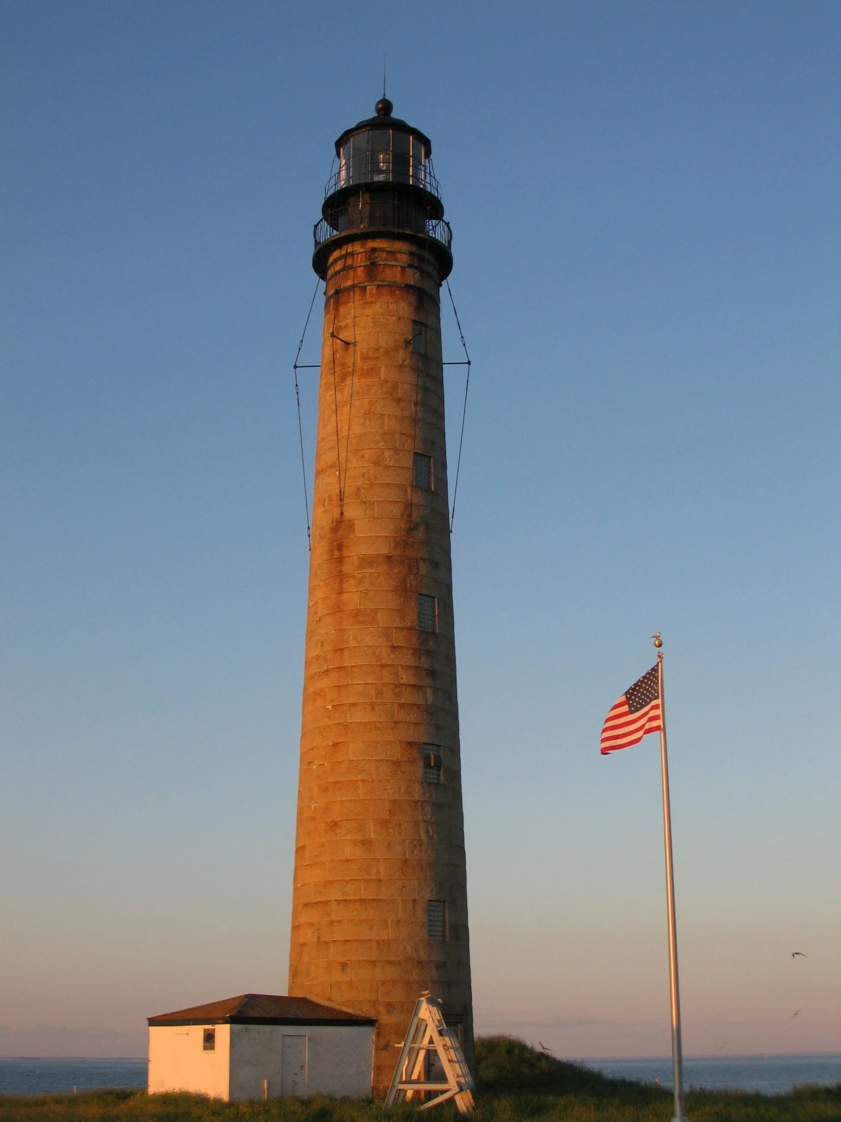A historic lighthouse standing tall with a small white building at its base, American flag flying nearby, with a clear blue sky and ocean in the background.