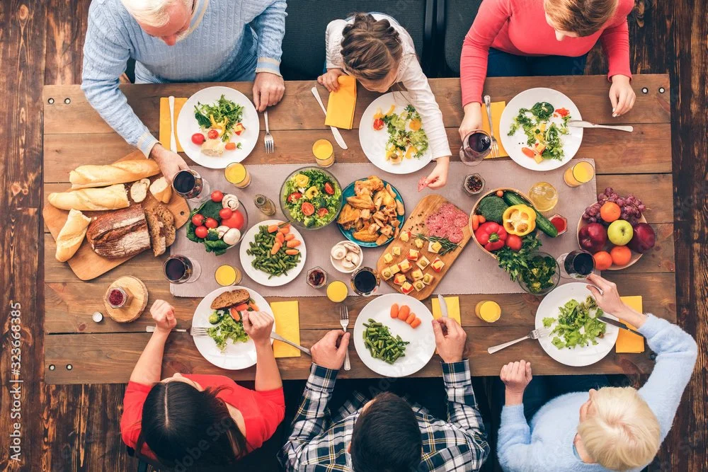 Family having dinner at a wooden table with salads, bread, vegetables, cheese, and drinks.