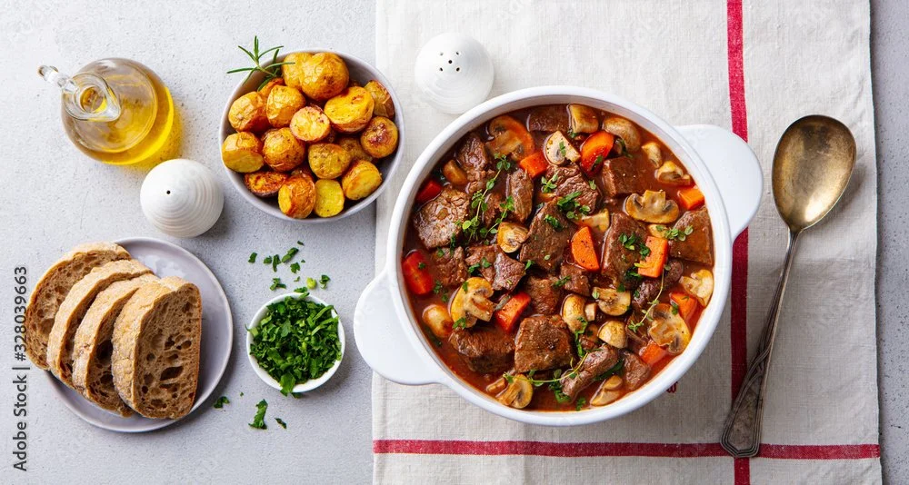 A bowl of beef stew with carrots and mushrooms, surrounded by slices of bread, roasted potatoes, chopped herbs, and condiments, on a white surface with a striped napkin.