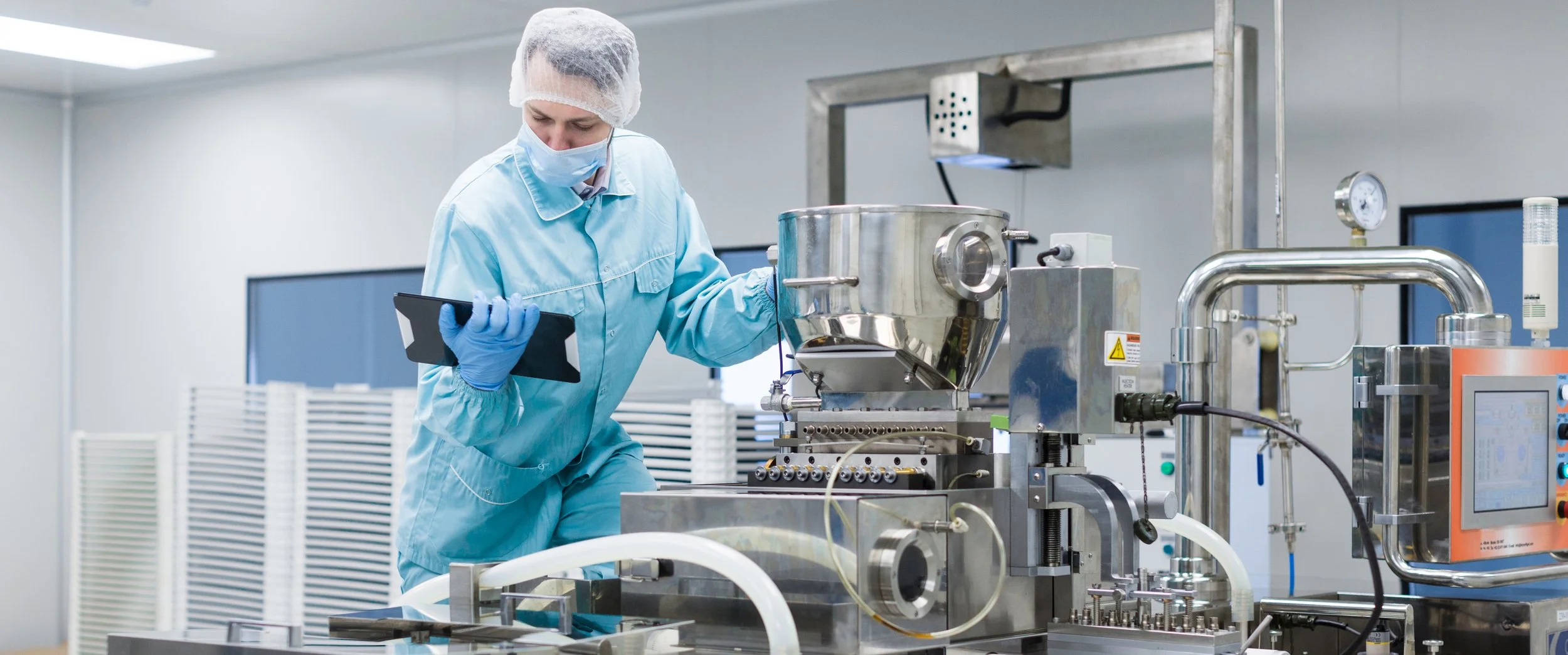 A food scientist in a laboratory operating stainless steel machinery, wearing a hairnet, face mask, gloves, and a light blue lab coat.