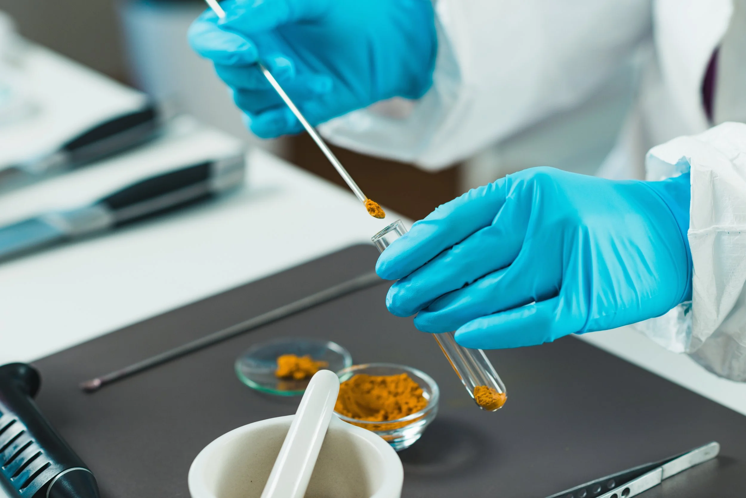 A food scientist wearing blue gloves and a white lab coat handling a test tube with spices, with other lab tools and spices on the table.