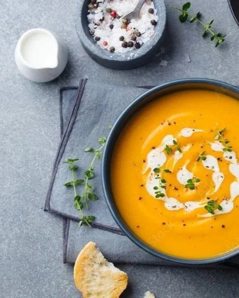 A bowl of butternut squash soup garnished with cream and microgreens, with a piece of bread on the side, salt and pepper in a grinder, and salt in a small jug.