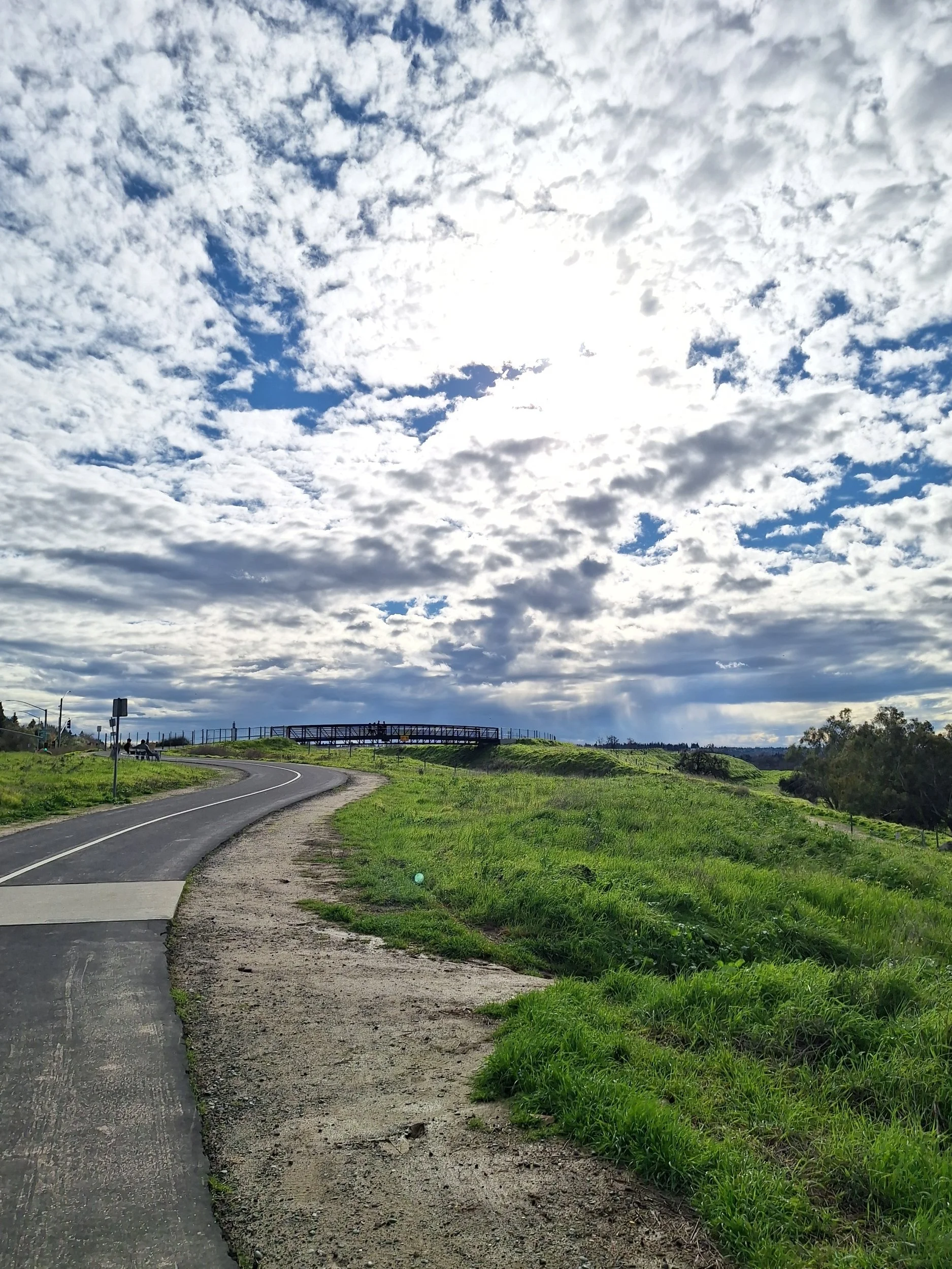 A winding road alongside a grassy field under a partly cloudy sky with the sun shining through the clouds.