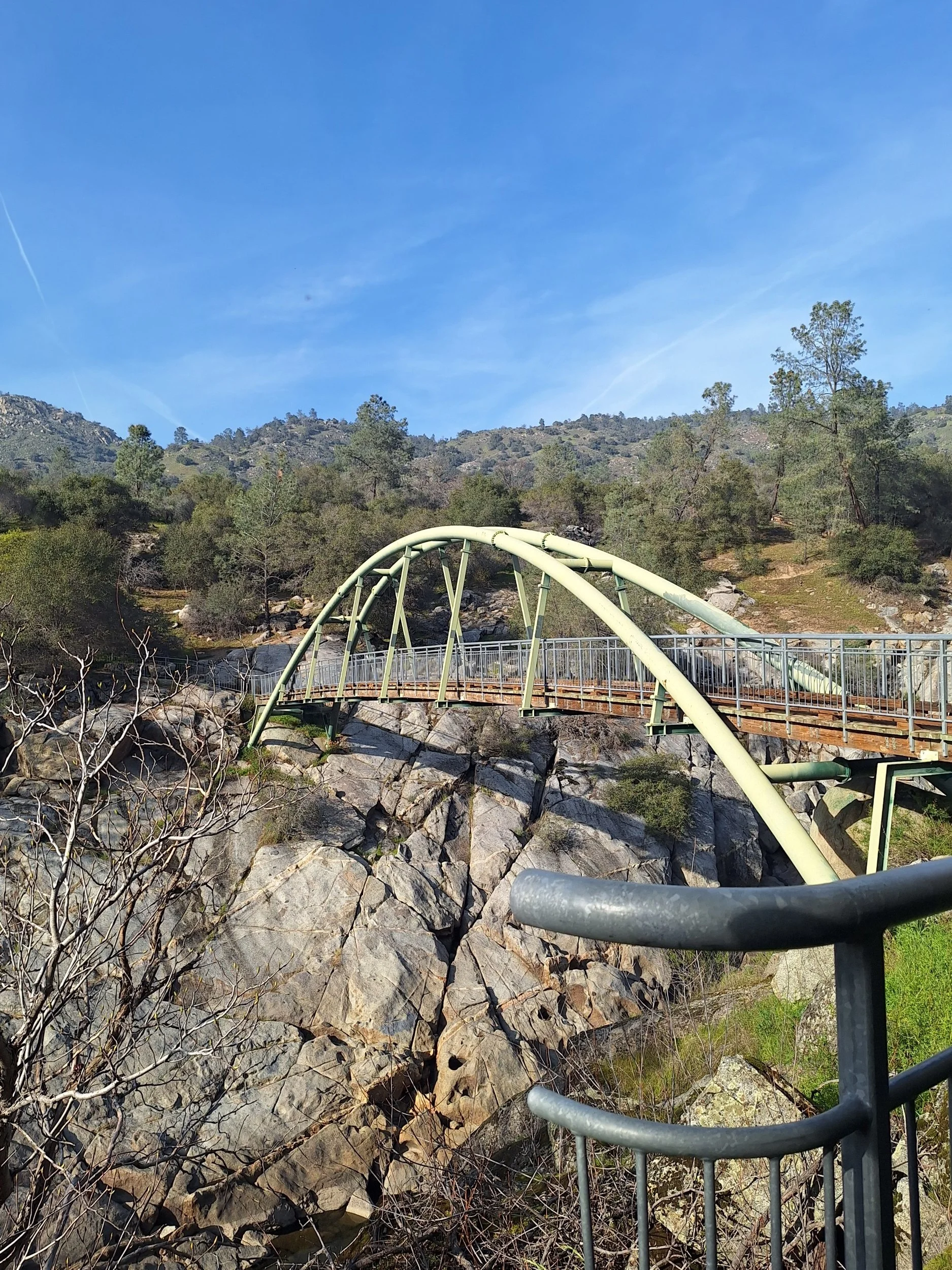 Steel arch and walkway bridge over rocky terrain with trees and mountains in the background under a blue sky.
