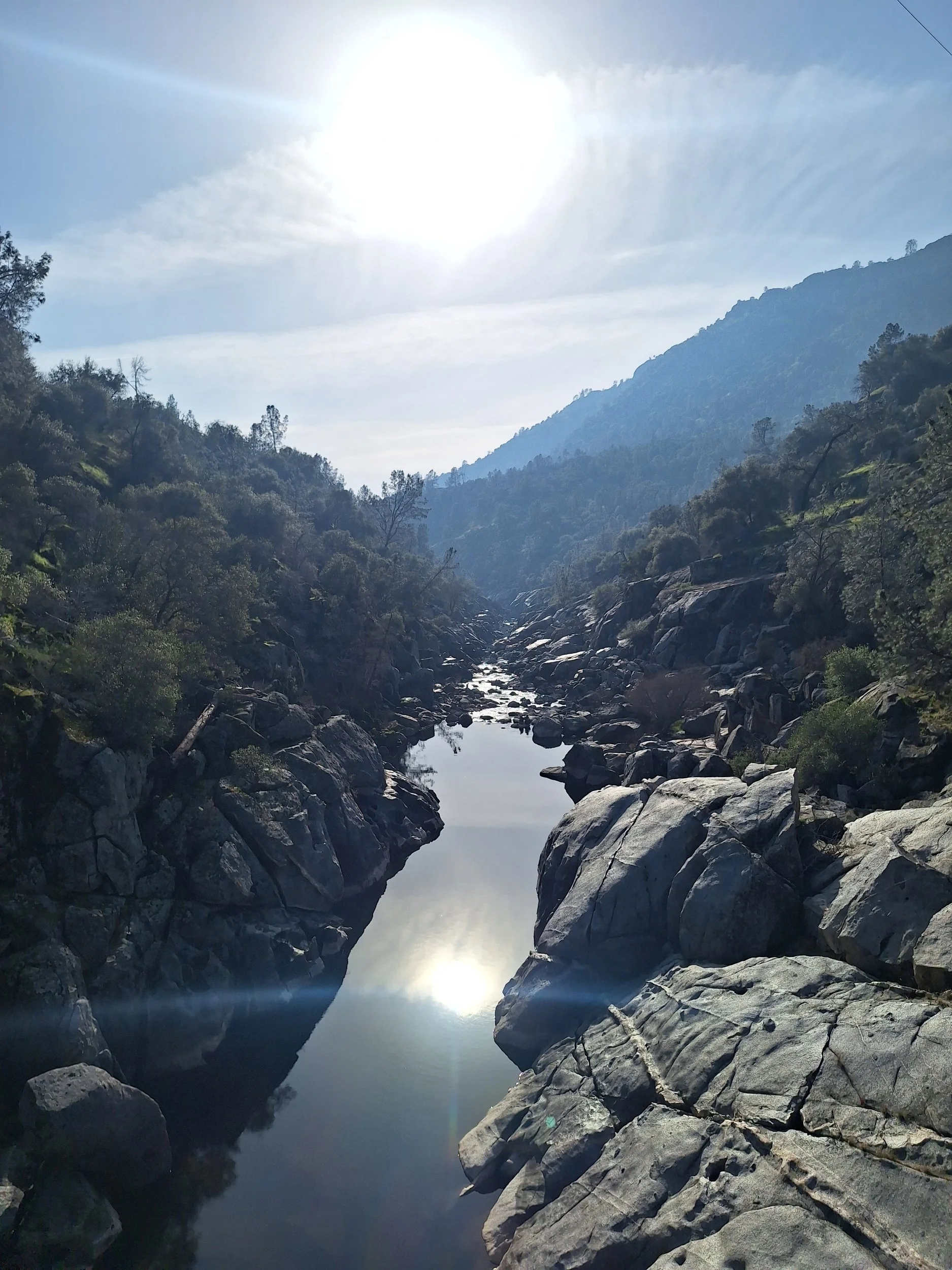 A river flowing through a rocky canyon with trees on both sides and mountains in the background, under a partly cloudy sky with the sun shining.