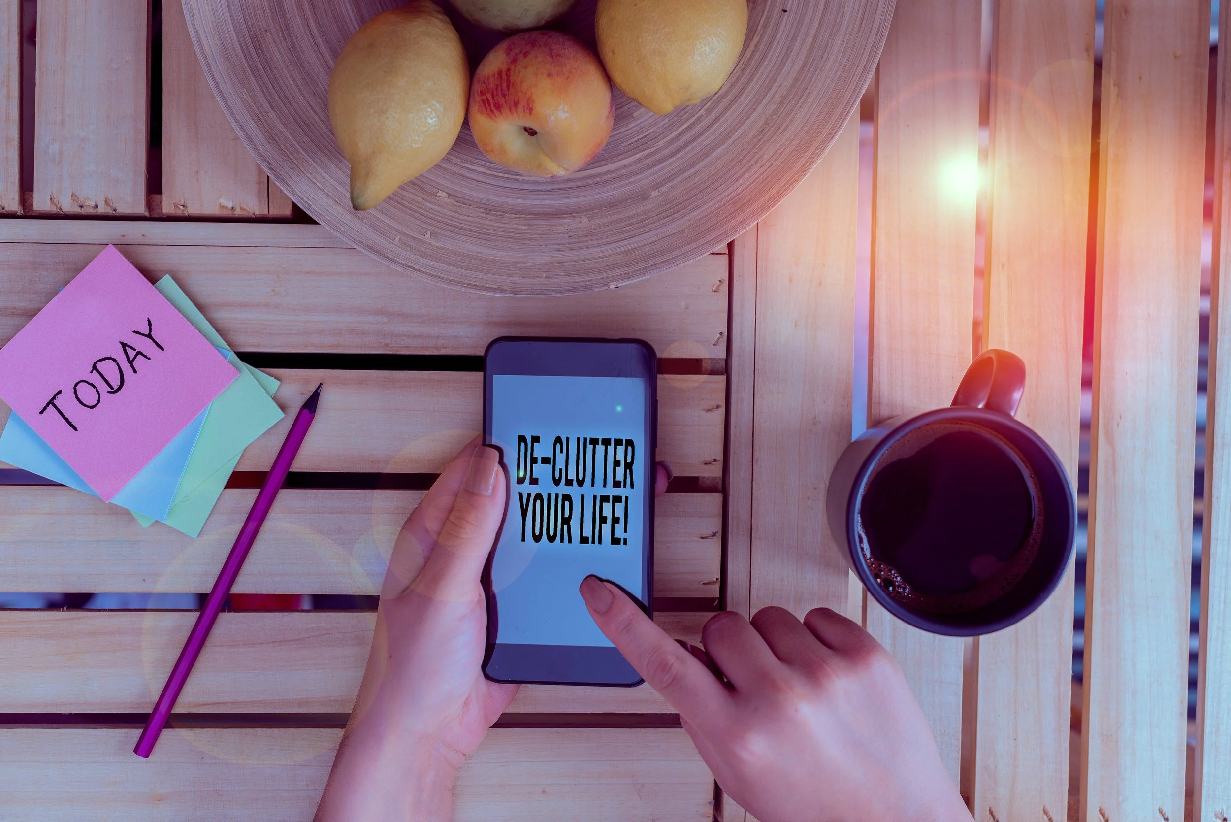 A person holding a smartphone with the message 'De-clutter your life!' on the screen, surrounded by a coffee mug, a bowl of pears and apples, sticky notes with 'Today' written on one, and a pink pen on a wooden table.