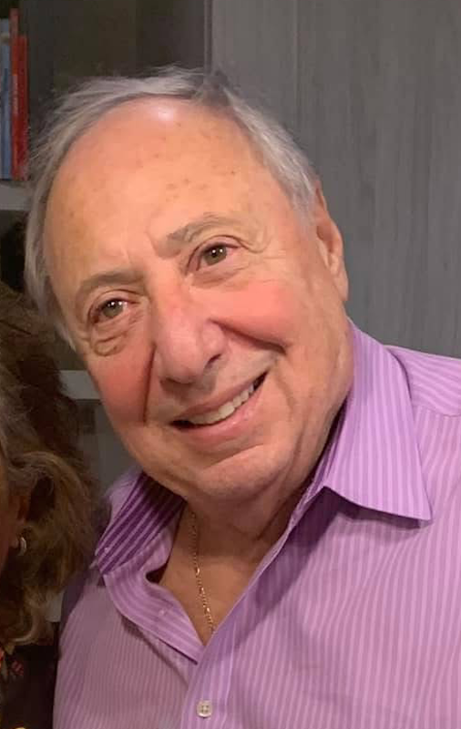 Close-up of an elderly man smiling, wearing a purple striped dress shirt and gold necklace, in an indoor setting.