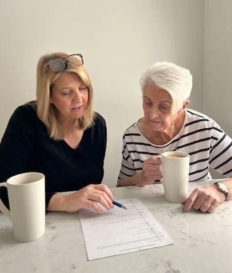 Two women sit at a table with documents and coffee mugs, discussing together.