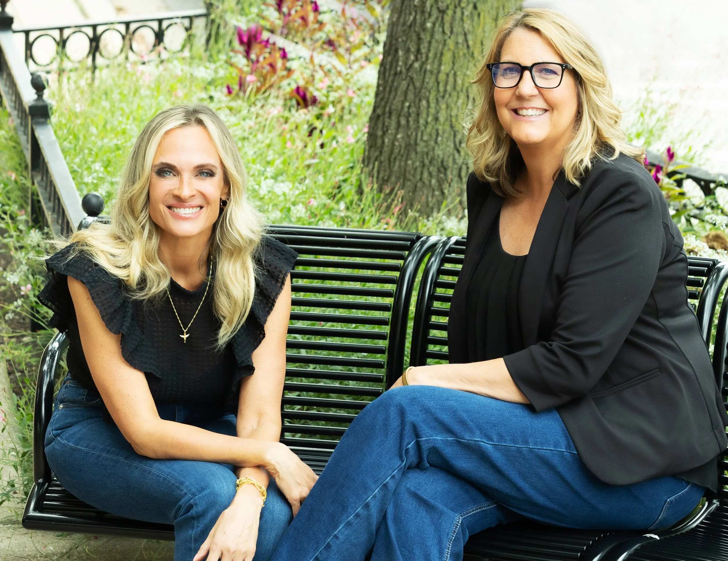 Two women sitting on a black metal bench outdoors, surrounded by greenery and flowers, smiling at the camera.