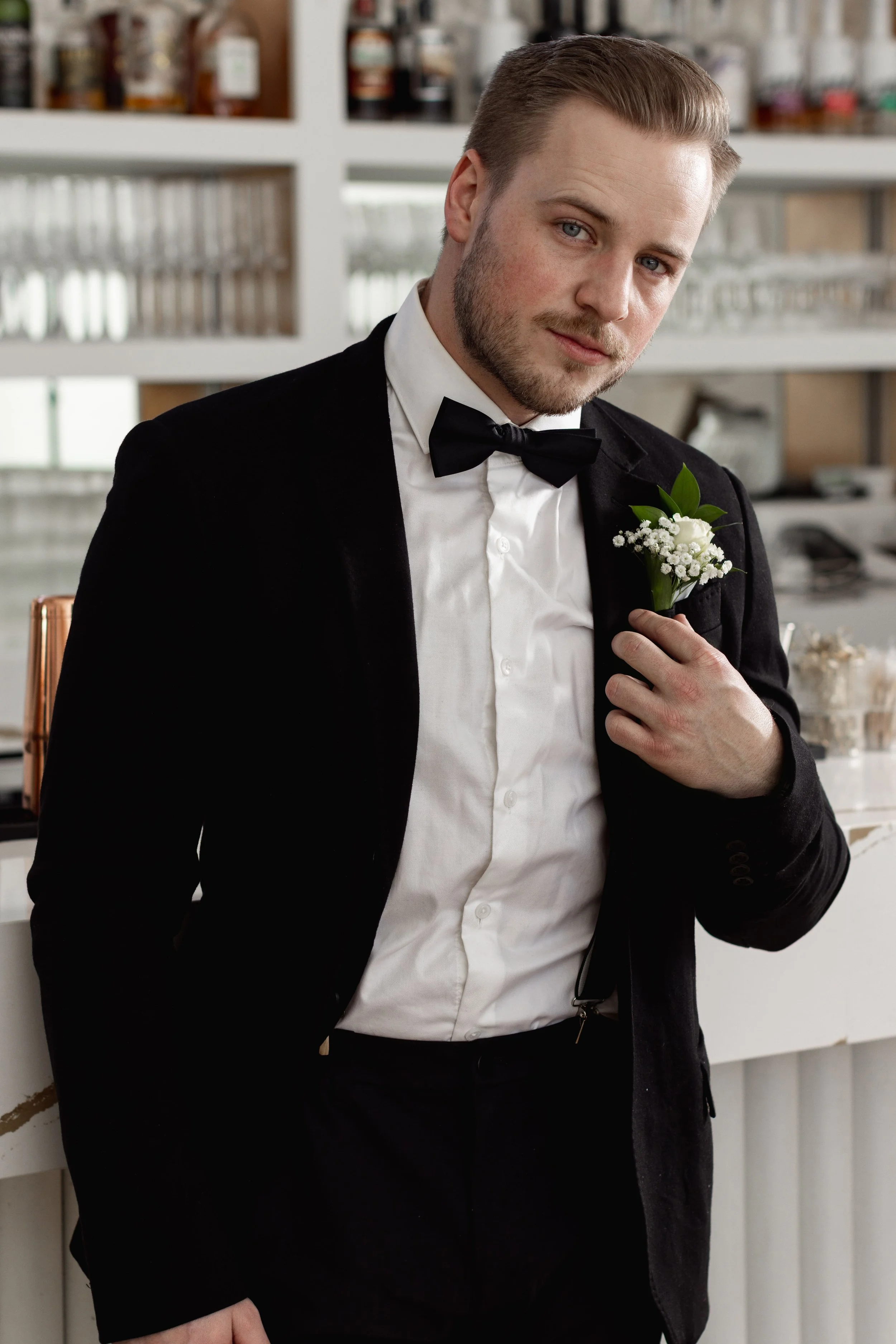 A man dressed in a tuxedo with a bow tie and a white flower boutonniere, posing indoors.