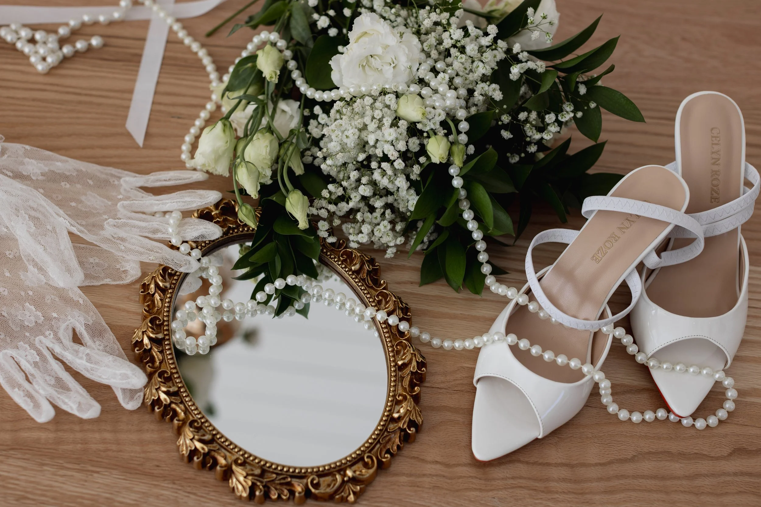 Wedding accessories including white high-heeled shoes, a bouquet of white flowers, a vintage mirror, pearl necklaces, a lace glove, and a pair of white earrings placed on a wooden surface.