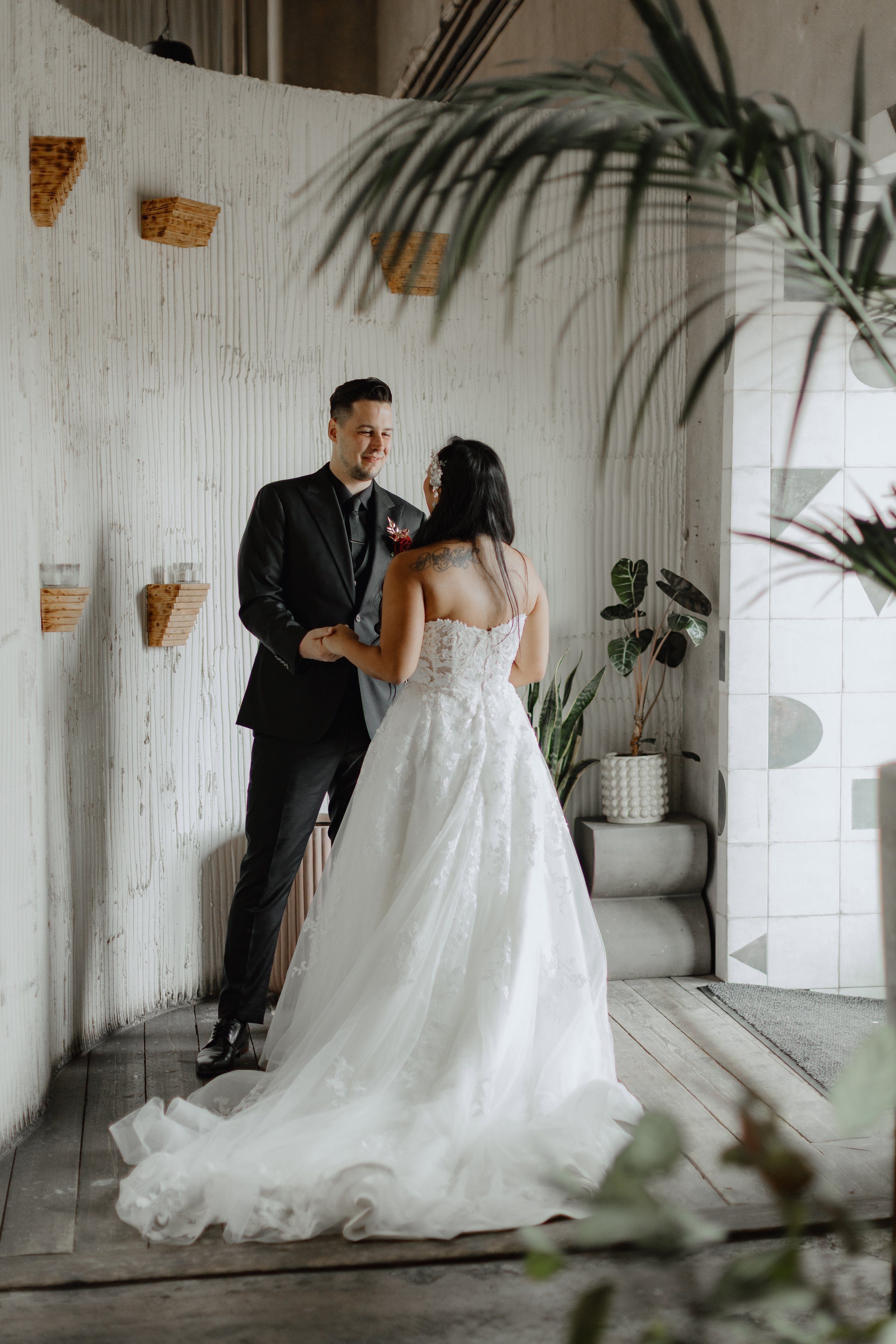 A bride and groom holding hands during their wedding ceremony indoors, with the groom in a black suit and the bride in a white lace wedding gown, surrounded by plants and a textured wall.