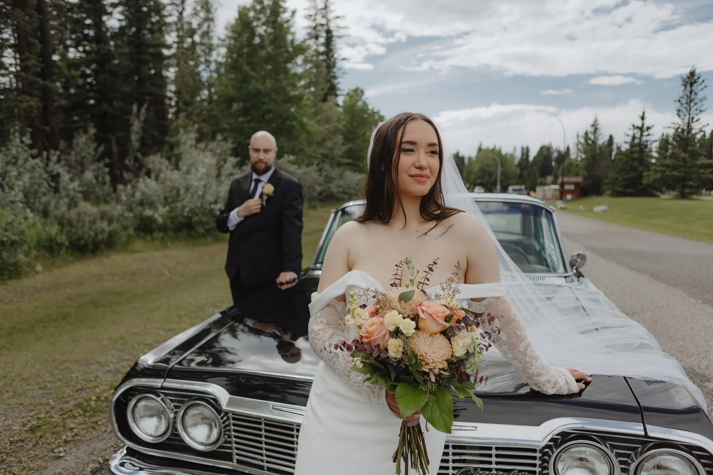 A bride in a white off-the-shoulder wedding dress holding a bouquet of peach, cream, and burgundy flowers, standing in front of a vintage black car with a veil, with a groom in a black suit and white shirt sitting on the hood, in a lush outdoor setting with trees and a cloudy sky.