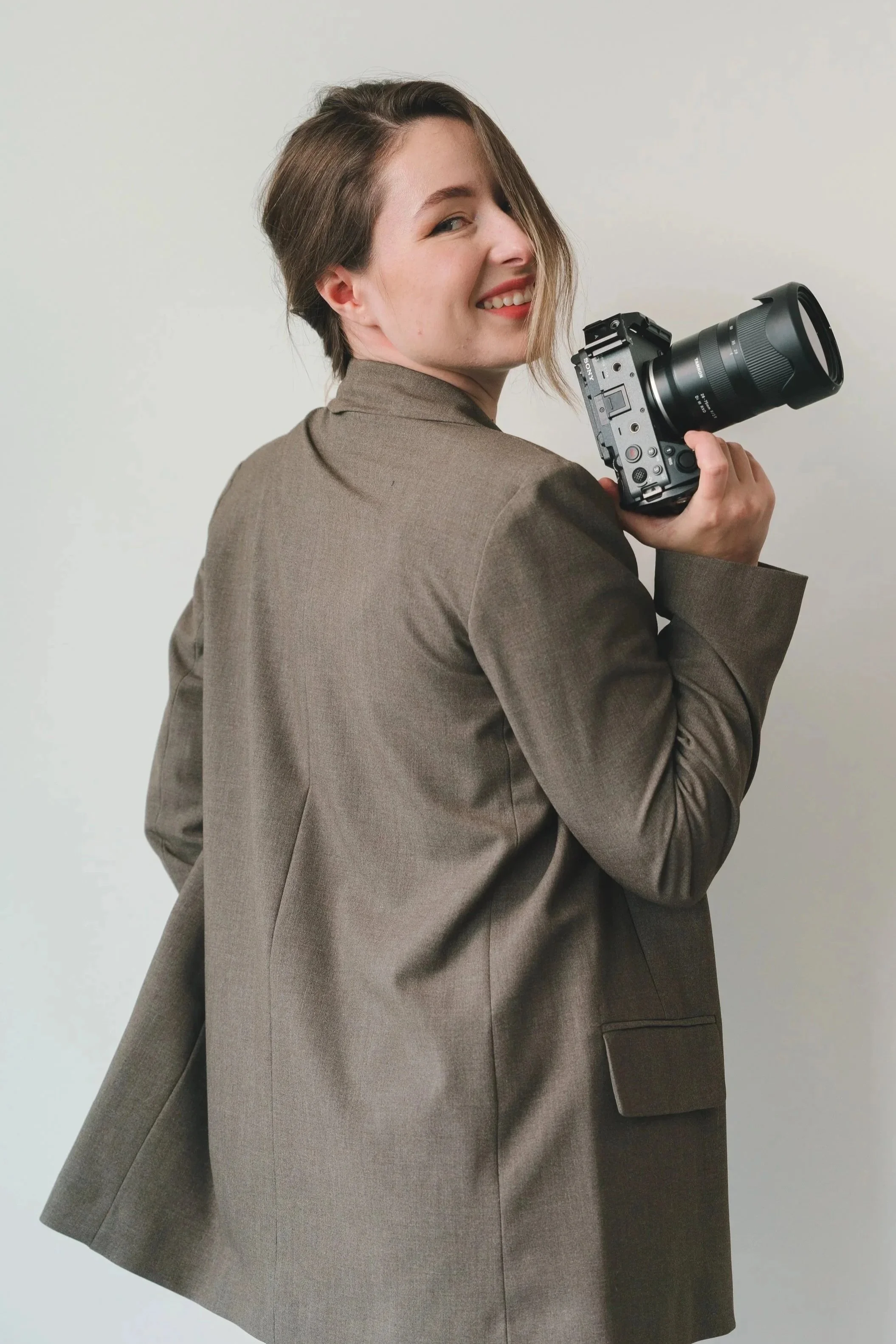 A woman with short brown hair, wearing a brown blazer, smiling and holding a professional camera.