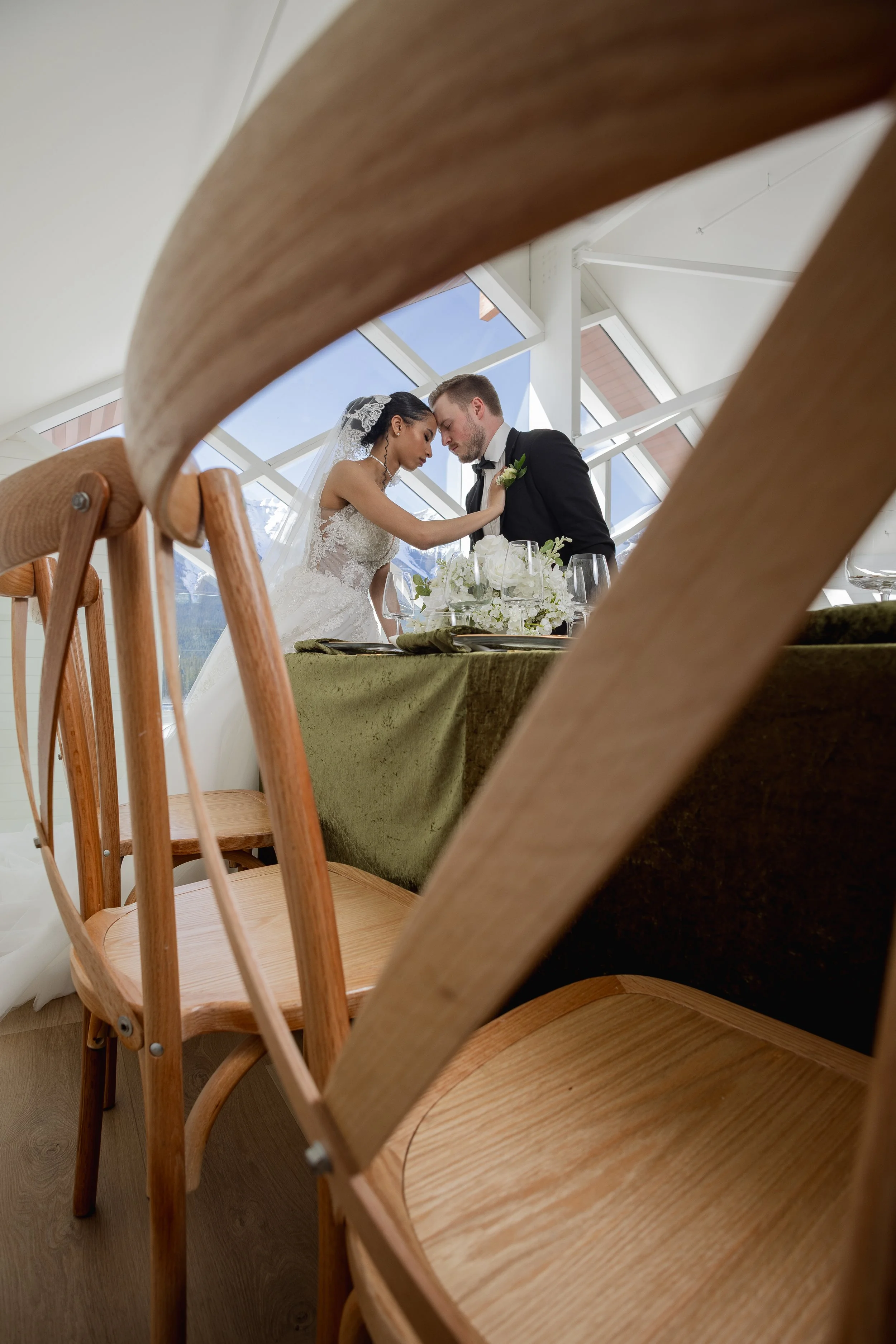 A bride and groom sharing an intimate moment at their wedding reception, seen through the gaps of wooden chairs surrounding the table.