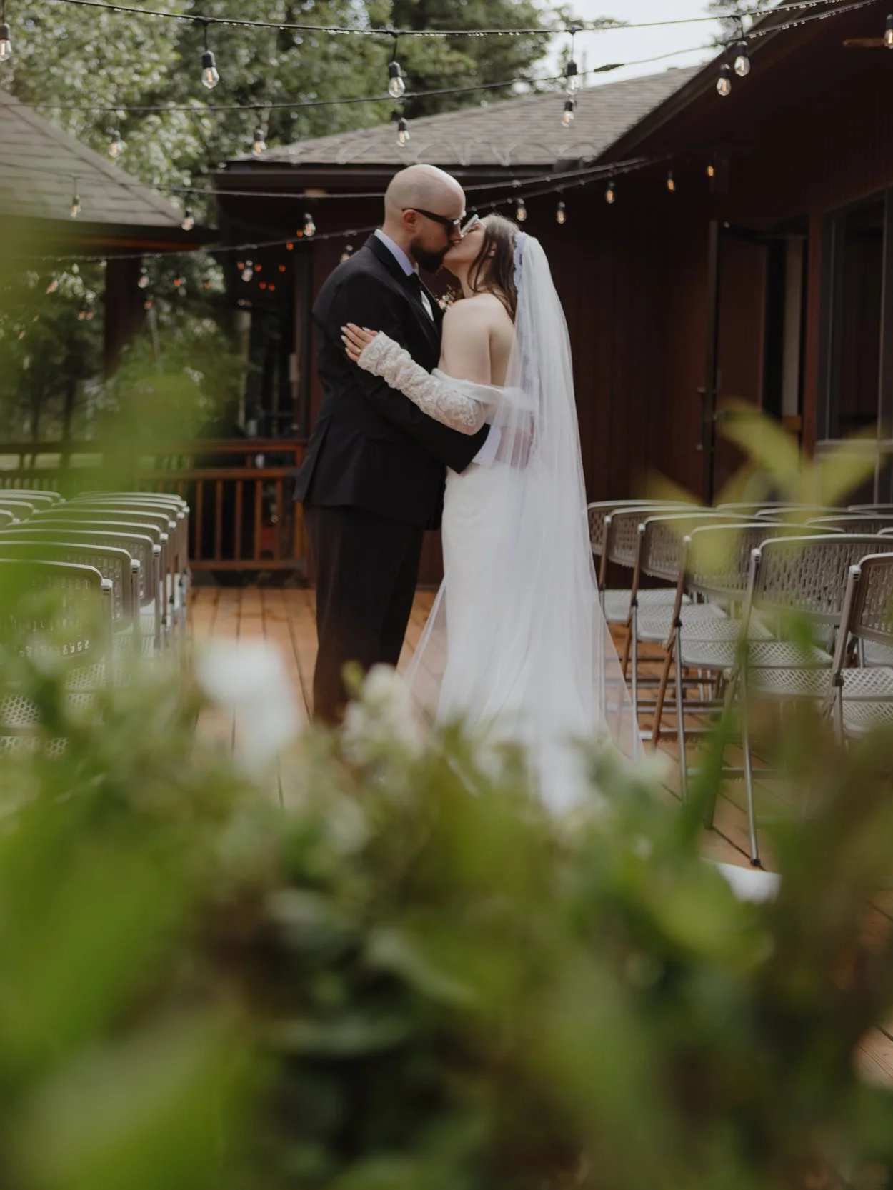 A wedding ceremony with a bride and groom sharing a kiss outdoors on a wooden deck, with string lights overhead and chairs lined up on either side.