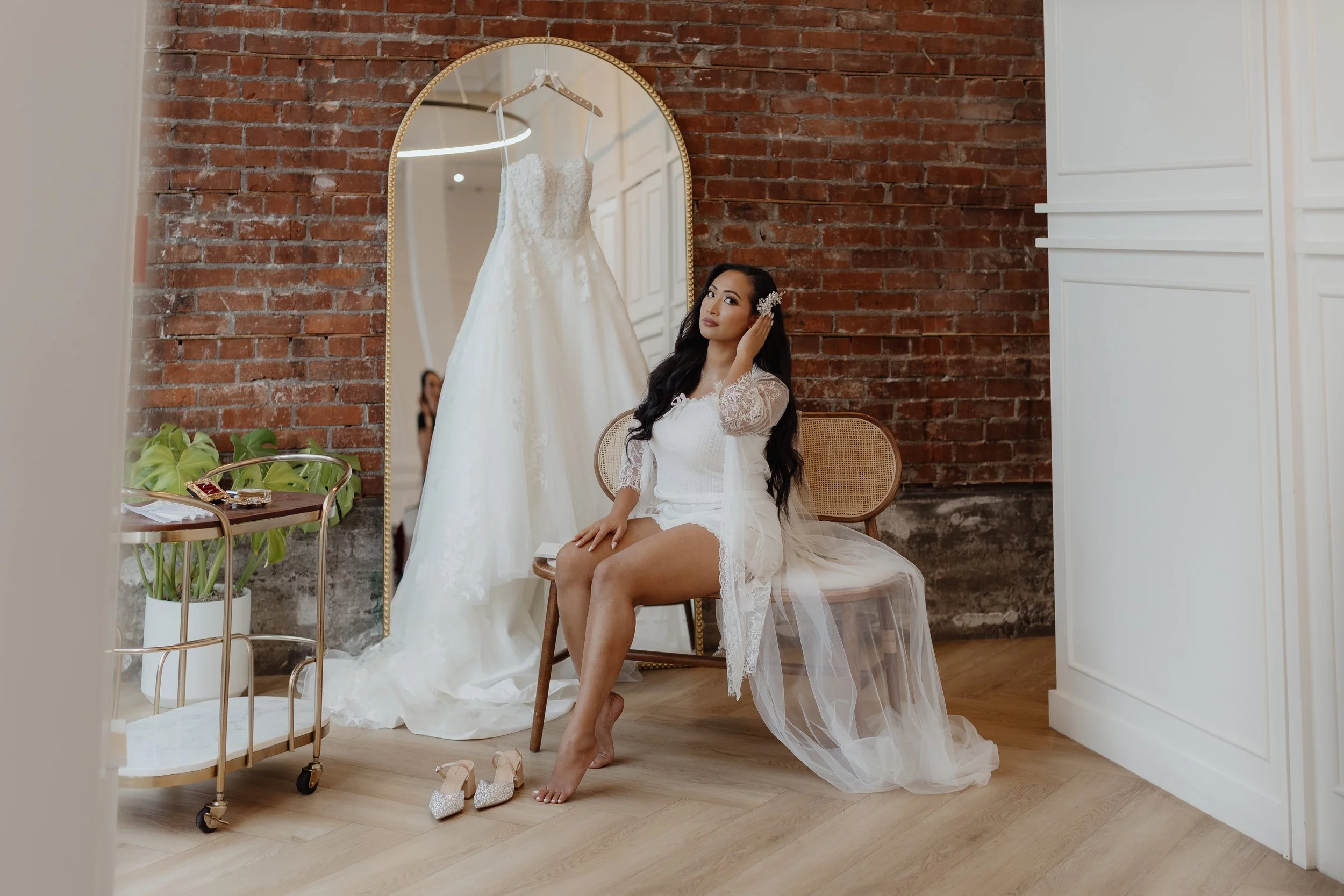 A woman in a white lace robe sits on a wooden chair in a room with exposed brick wall, looking at herself in a large mirror. Behind her, a white wedding dress hangs on the wall, and she is touching her hair. On the floor, her sparkly high heels are visible, and to her left, a gold cart with green plants and small items.