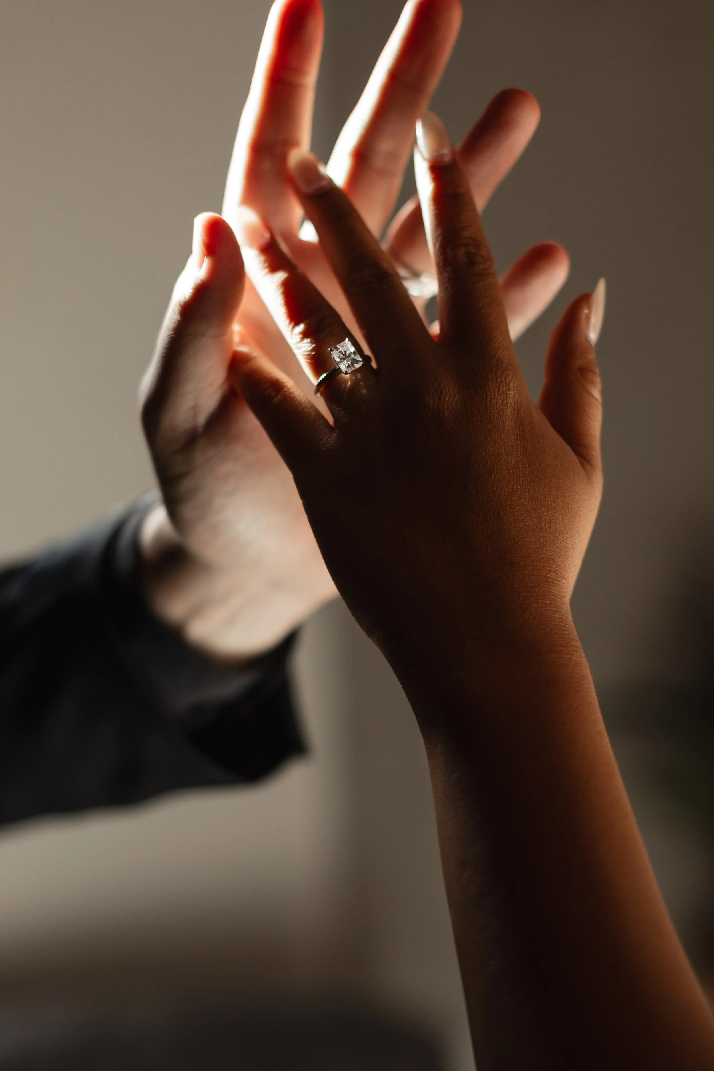Close-up of two hands, one darker-skinned and one lighter-skinned, touching. The darker-skinned hand has a large diamond ring on the ring finger. The background is blurred and the lighting highlights the hands and ring.