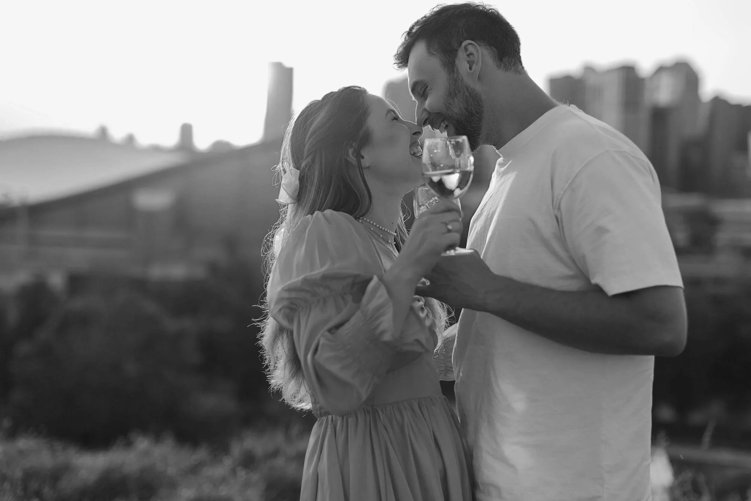 A couple smiling and enjoying wine, standing close together outdoors in a cityscape, sharing an intimate moment.